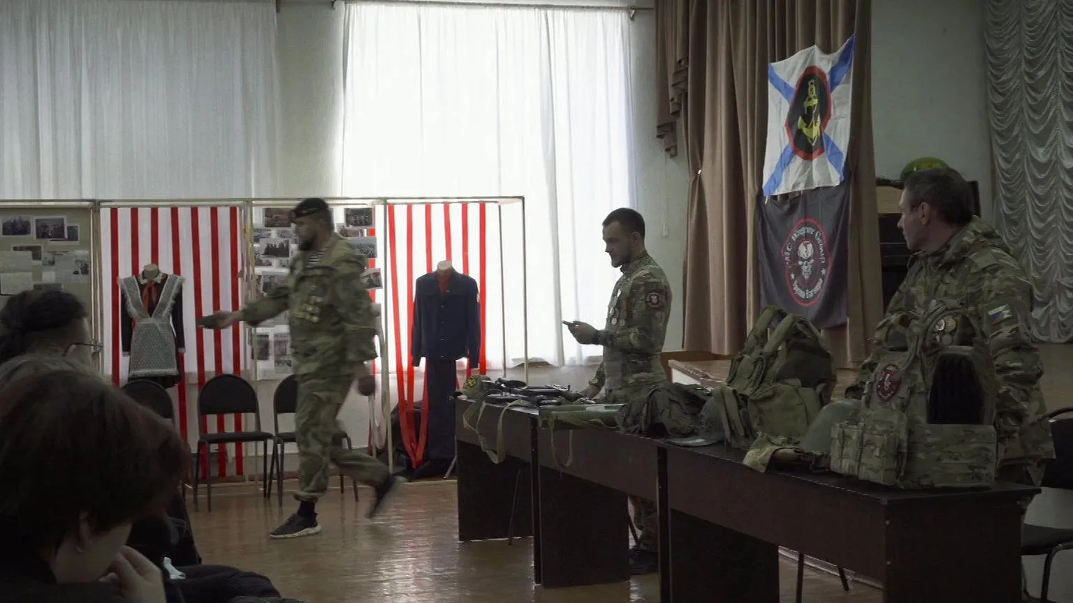 Pavel Talankin/BBC Storyville/Made in Copenhagen Three wagner mercenaries in green camouflage. Two by the stage, the third going towards pupils to show a mine to them. On the left by the window is a woman's uniform