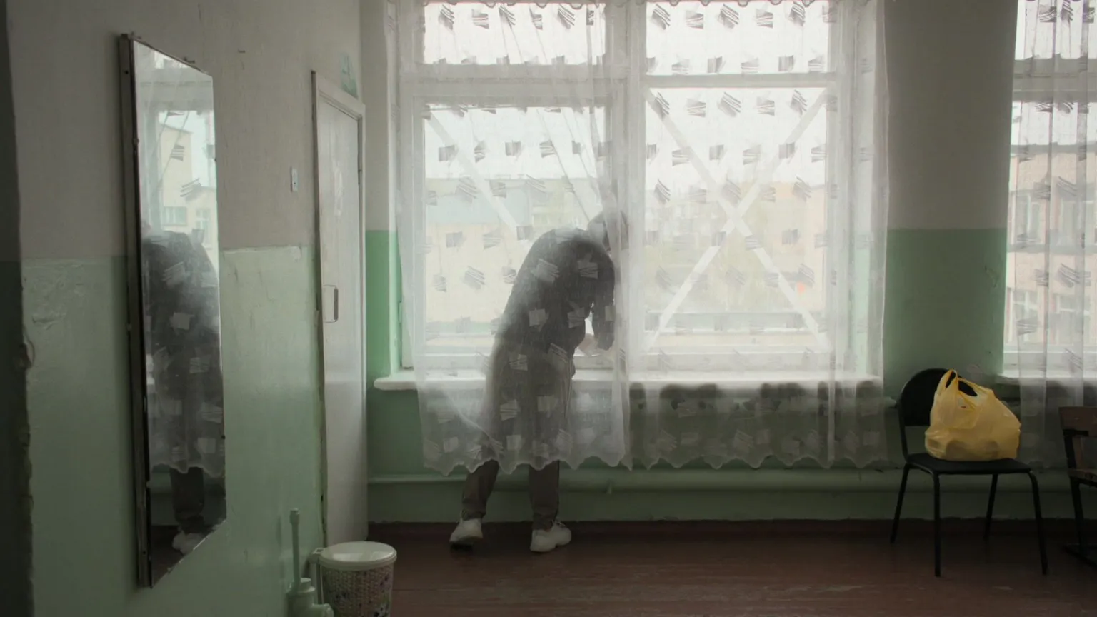 Pavel Talankin/BBC Storyville/Made in Copenhagen Pasha behind a net curtain sticking an X symbol on the window. chair with yellow bag. On left hand side a mirror. Green walls