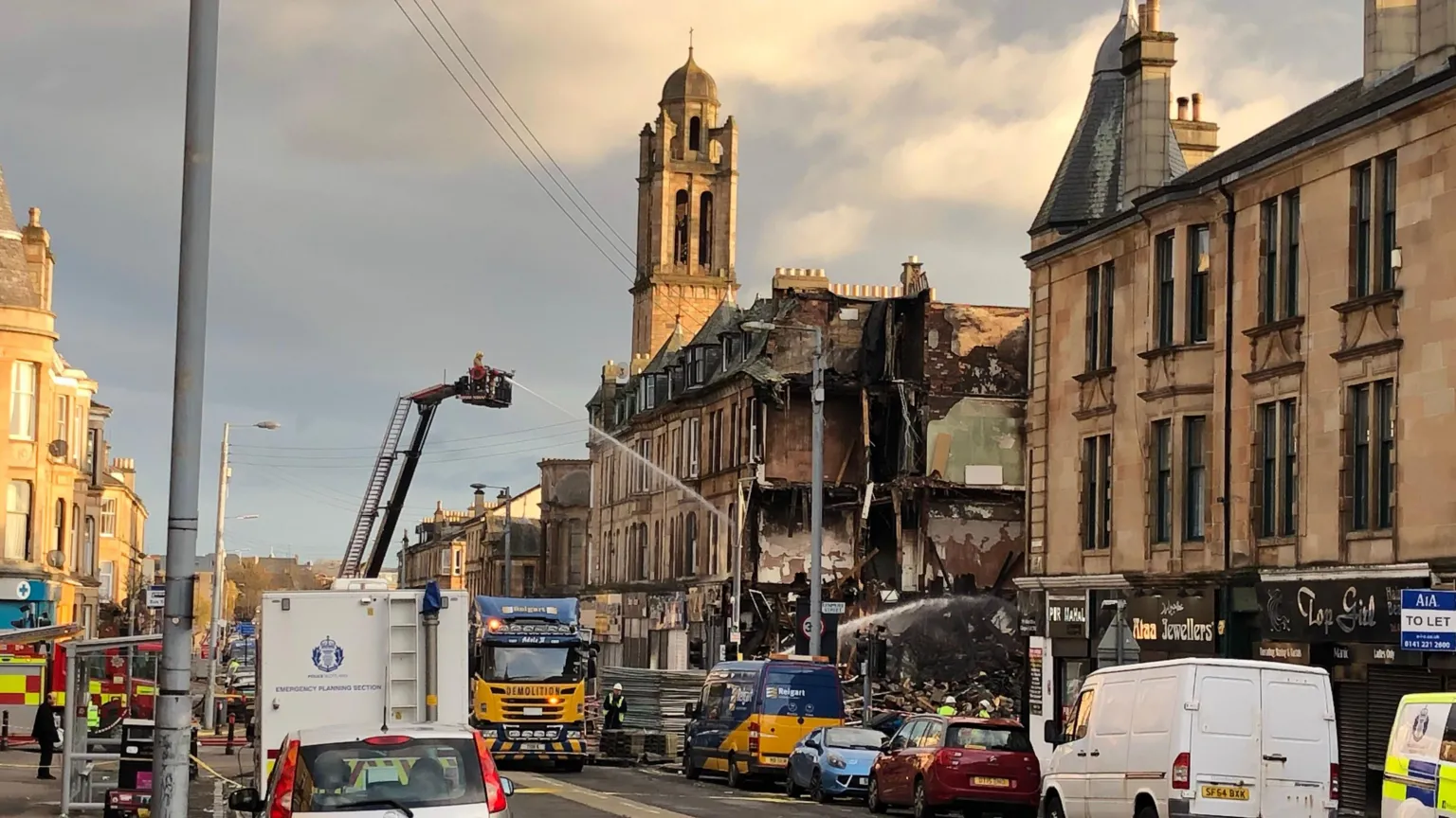 Fire-damaged tenement building. Firefighters are shooting water into a gap in the building. There is a large emergency service presence.