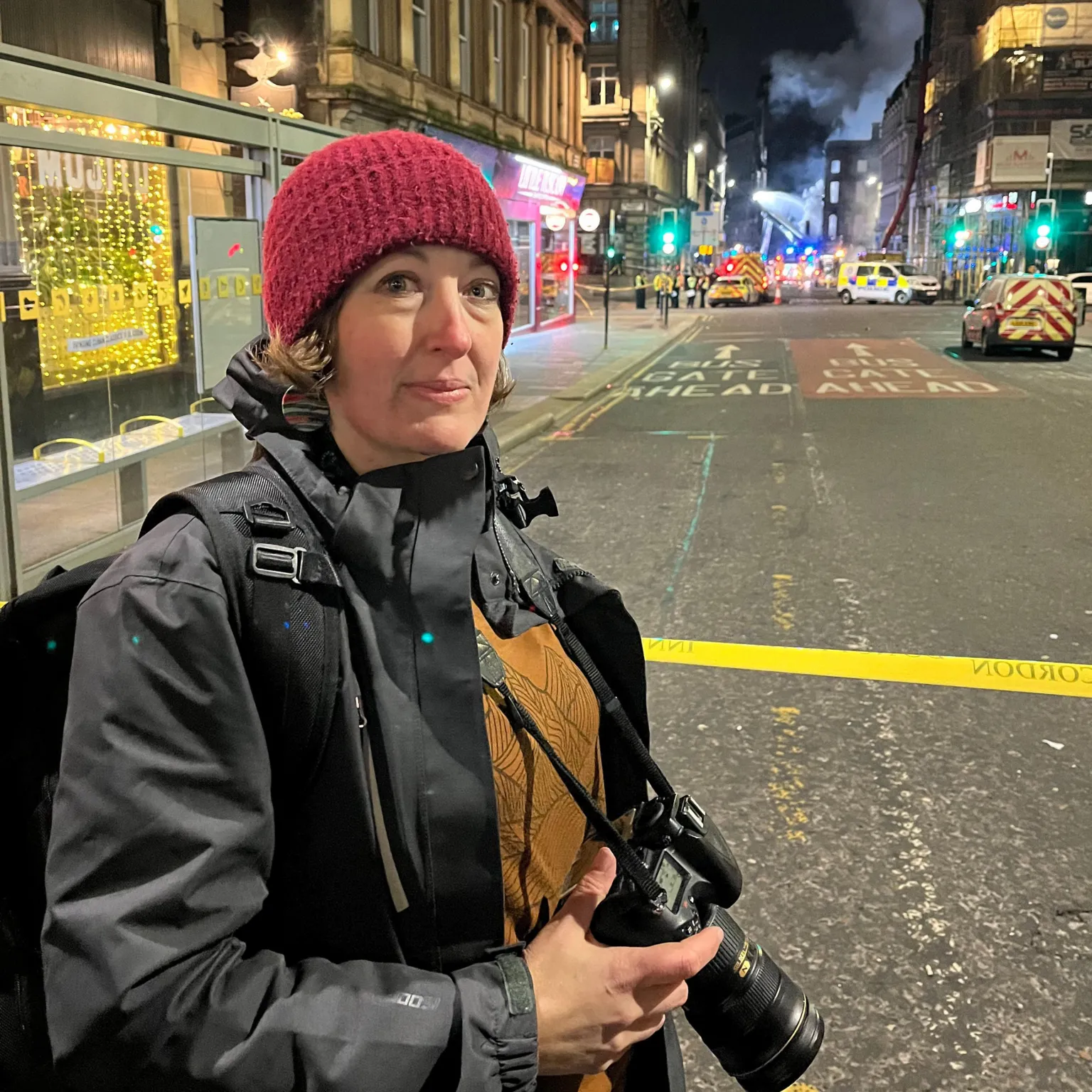 Michelel Cohen A woman in a red wollen hat and a black waterproof jacket. She has a camera hanging around her neck. She is standing in a city road and there is an emergency response to a fire in a building in the background.