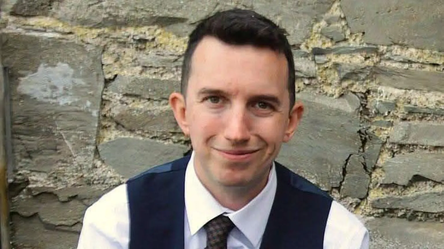 Andrew McCusker Andrew McCusker has short dark brown hair and is wearing a white shirt, dark tie and dark waistcoat. He is smiling in the picture Behind him is a stone wall.