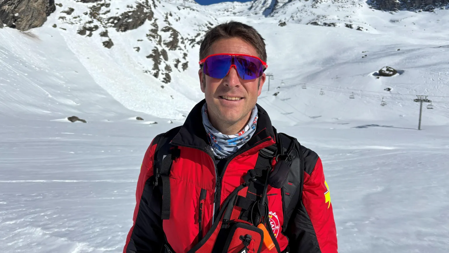 BBC/Paul Pradier A man is seen smiling wearing sunglasses and a red emergency services jacket against a background of a ski area.