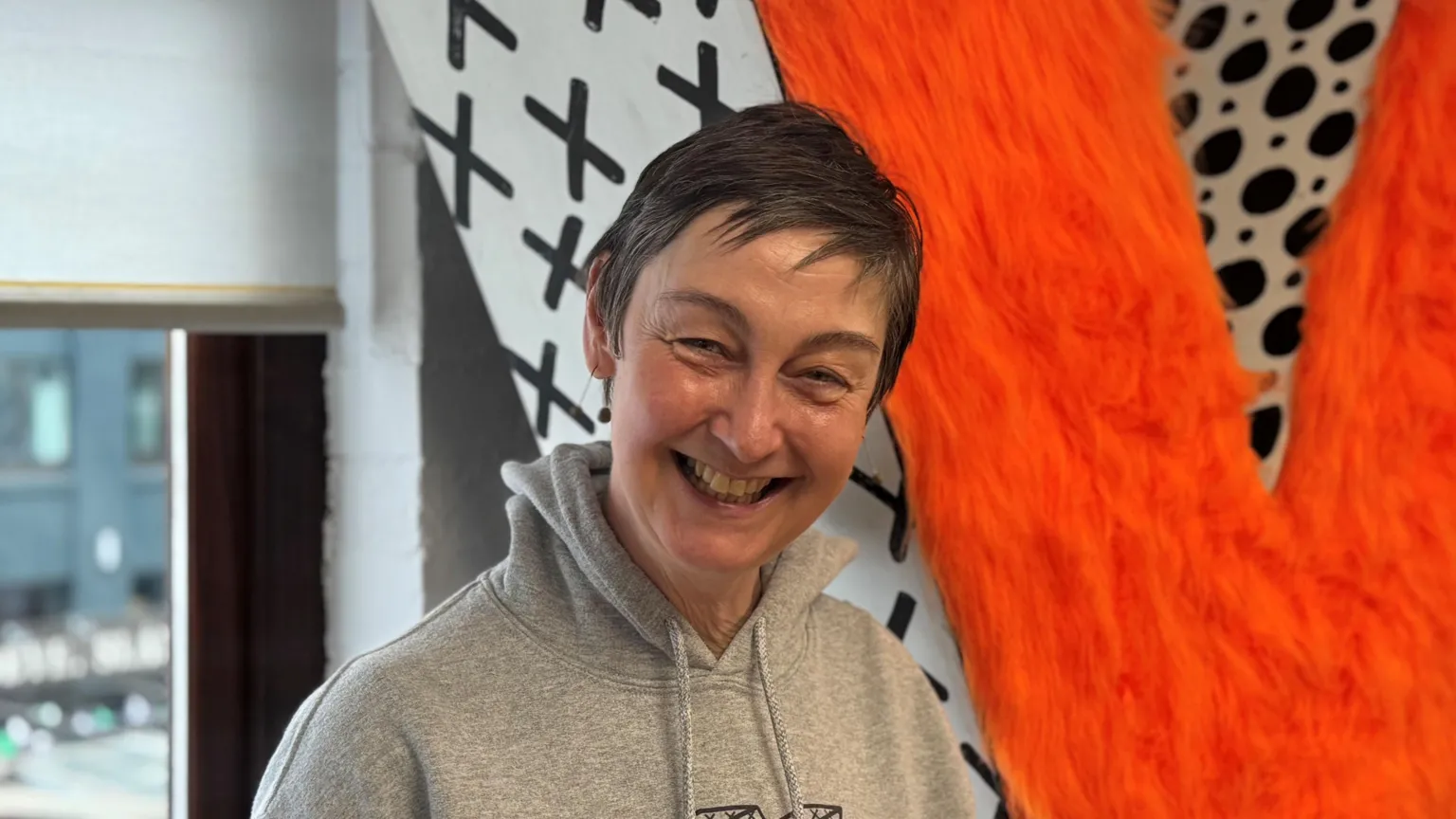 Woman wearing grey hoodie who has short brown hair is smiling at camera. There is a big neon orange letter Y behind her which is a piece of artwork.