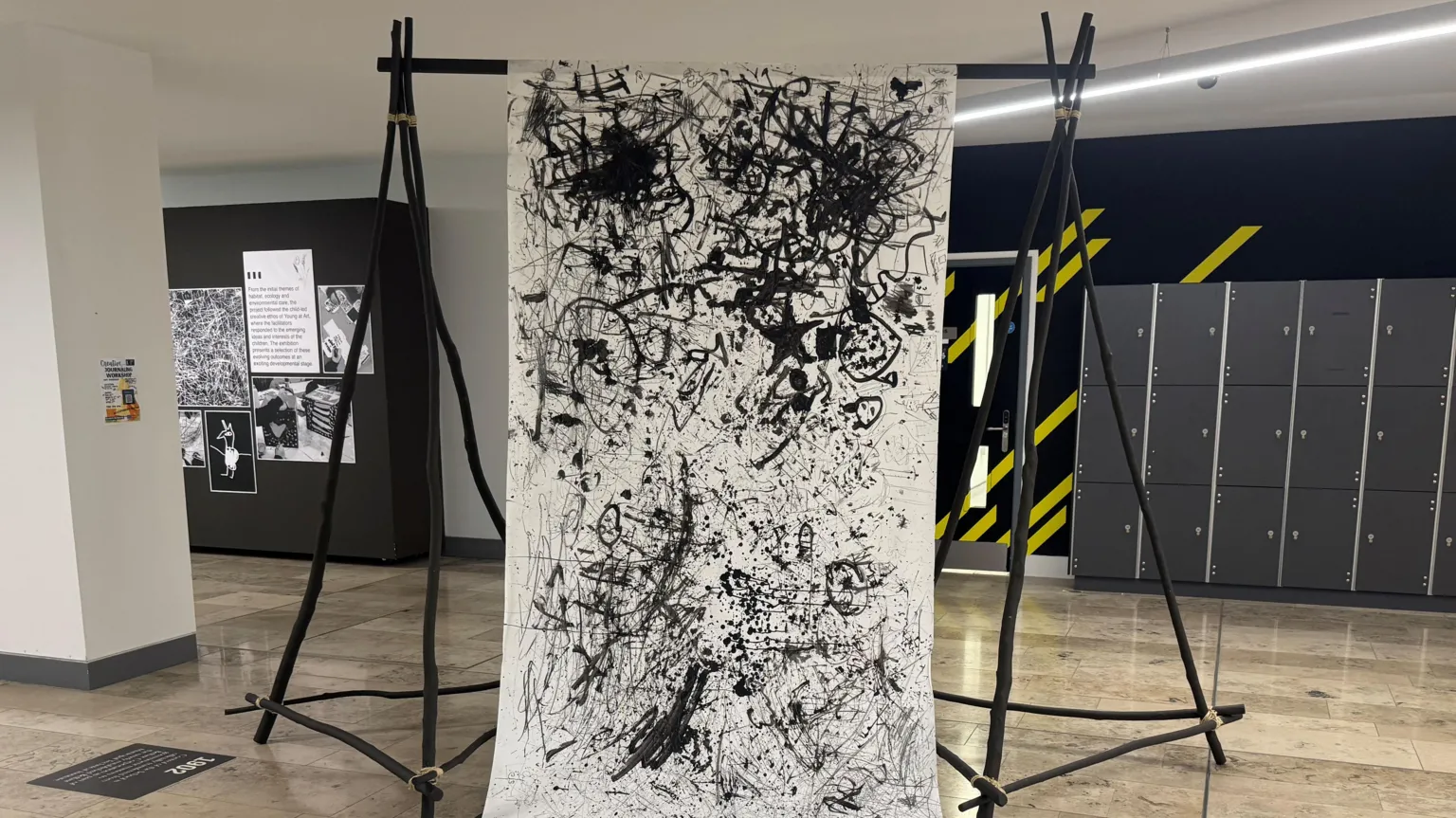 A large scroll of white paper is held up by black wooden beams. The children's art work is showcased on the scroll. The artwork is very abstract, lots of black lines and scribbles. 