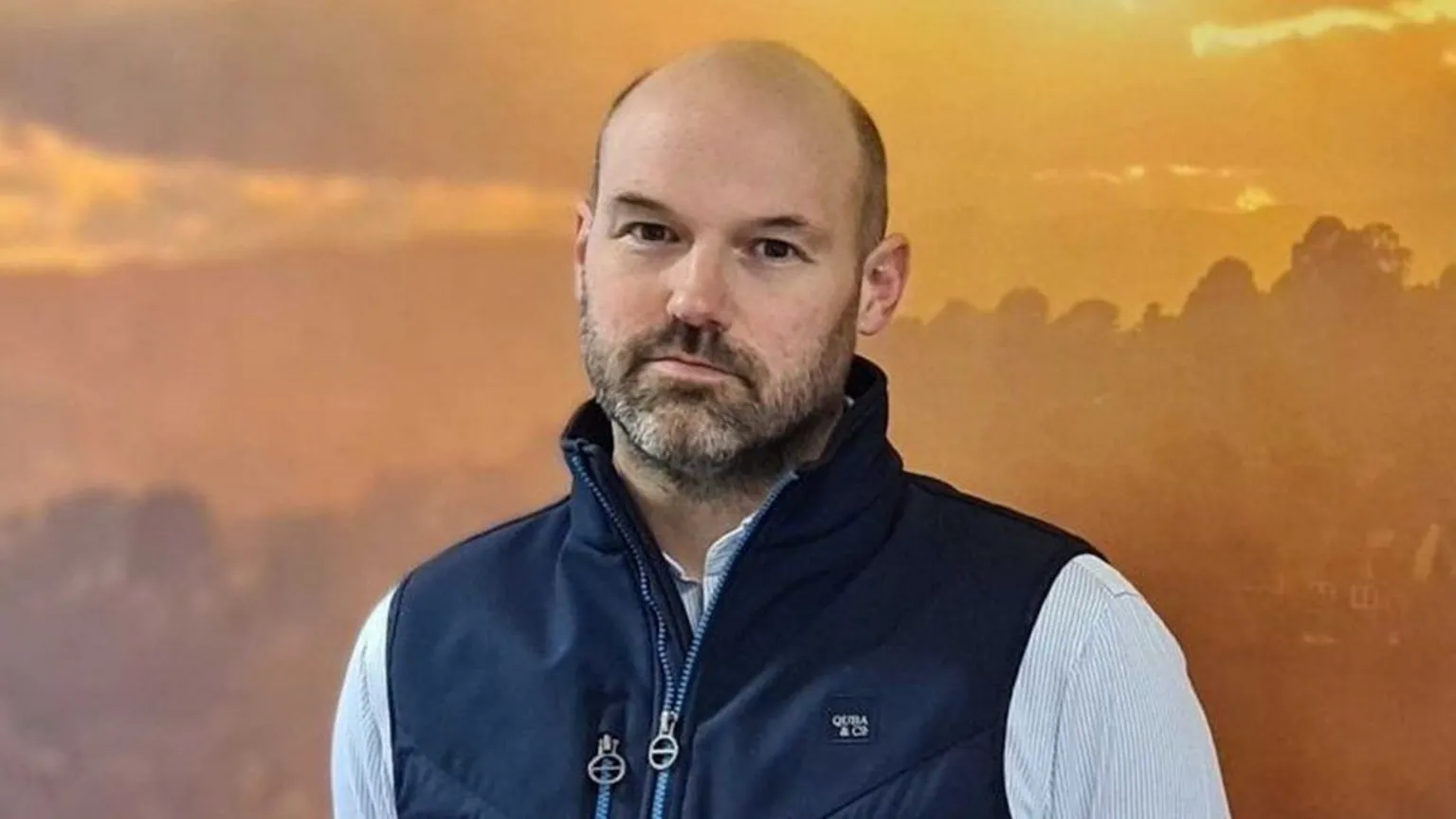 Tim Buckman Tim Buckman is wearing a navy bodywarmer over a white shirt with pale blue stripes. He has a beard and a shaved head. He is standing in front of a background of trees at either sunset or sunrise.