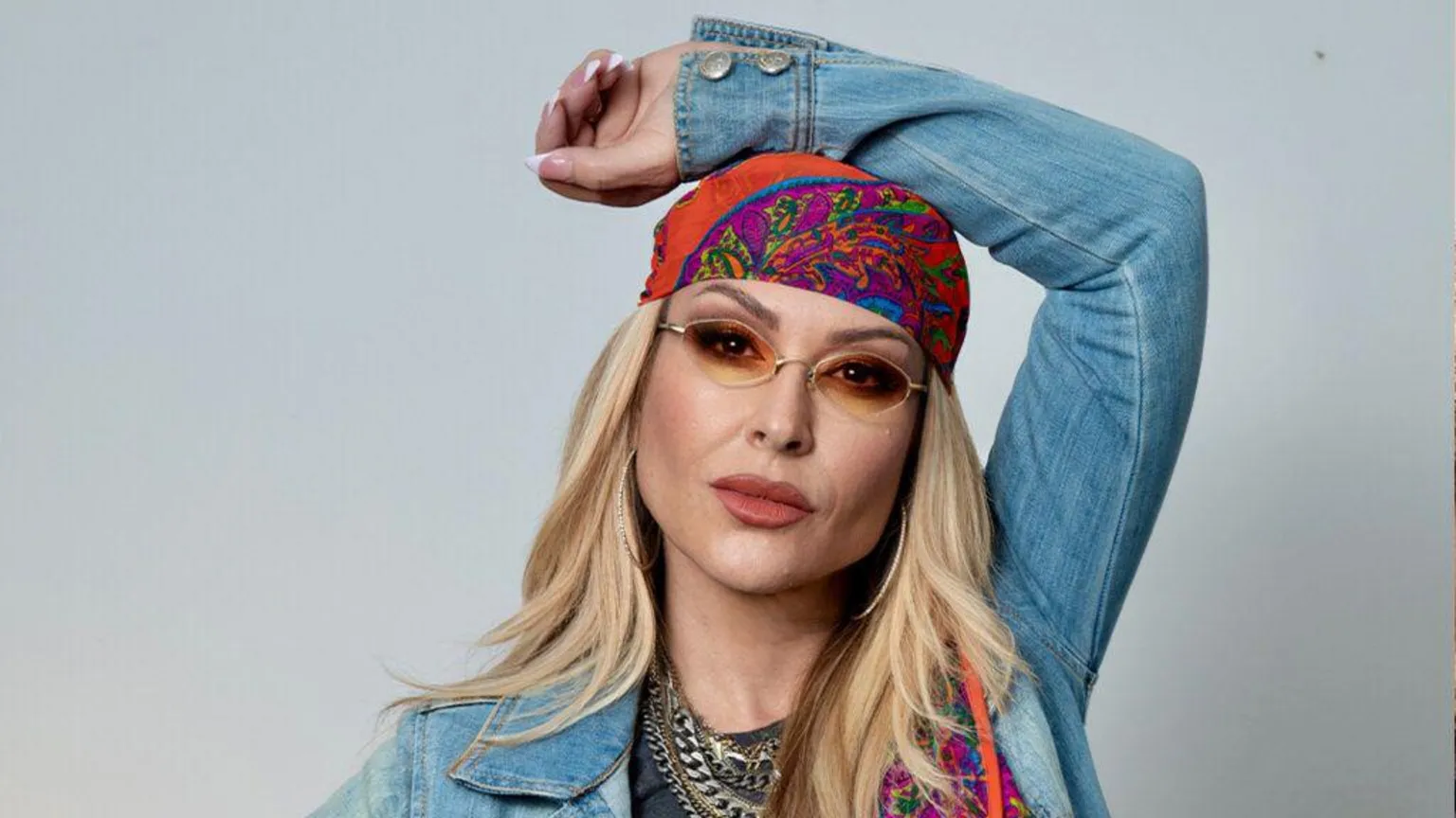 BBC/World of Wonder Photo of signer Anastacia posing with her hand above her head. She has long blonde hair and is wearing a denim jacket, bandana and sunglasses. 
