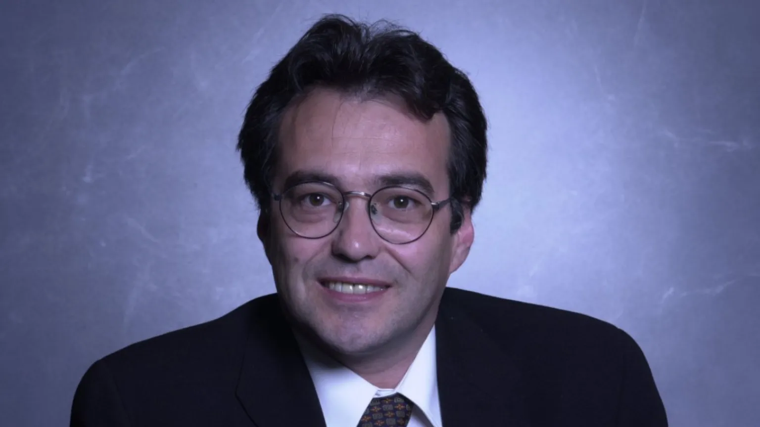 Phil Woolas, wearing a dark suit, tie and glasses, smiles straight in a posed photo from his time as an MP.