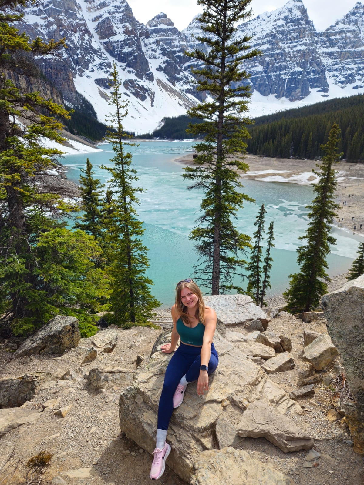 Jelena sitting on rocks with a vew of tall trees, a clear blue lake and snowy mountains behind