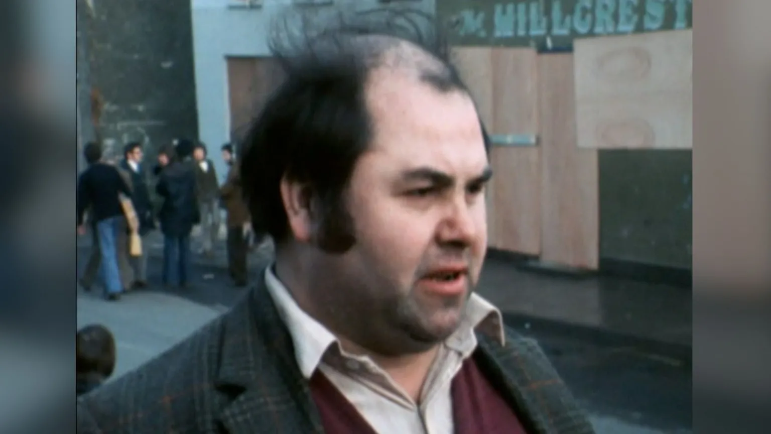 Archive footage shows a man with black hair long side burns and a tweed jacket. he's looking off camera talking to the reporter.