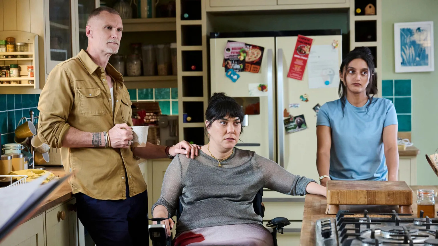 BBC / Roughcut TV / Adam Lawrence In a kitchen, a man holding a mug is wearing a yellow shirt and blue trousers, he stands next to a woman who uses a wheelchair, she is wearing a grey top and purple skirt. Another woman wearing a blue top stands to the right, in front of a chopping board.