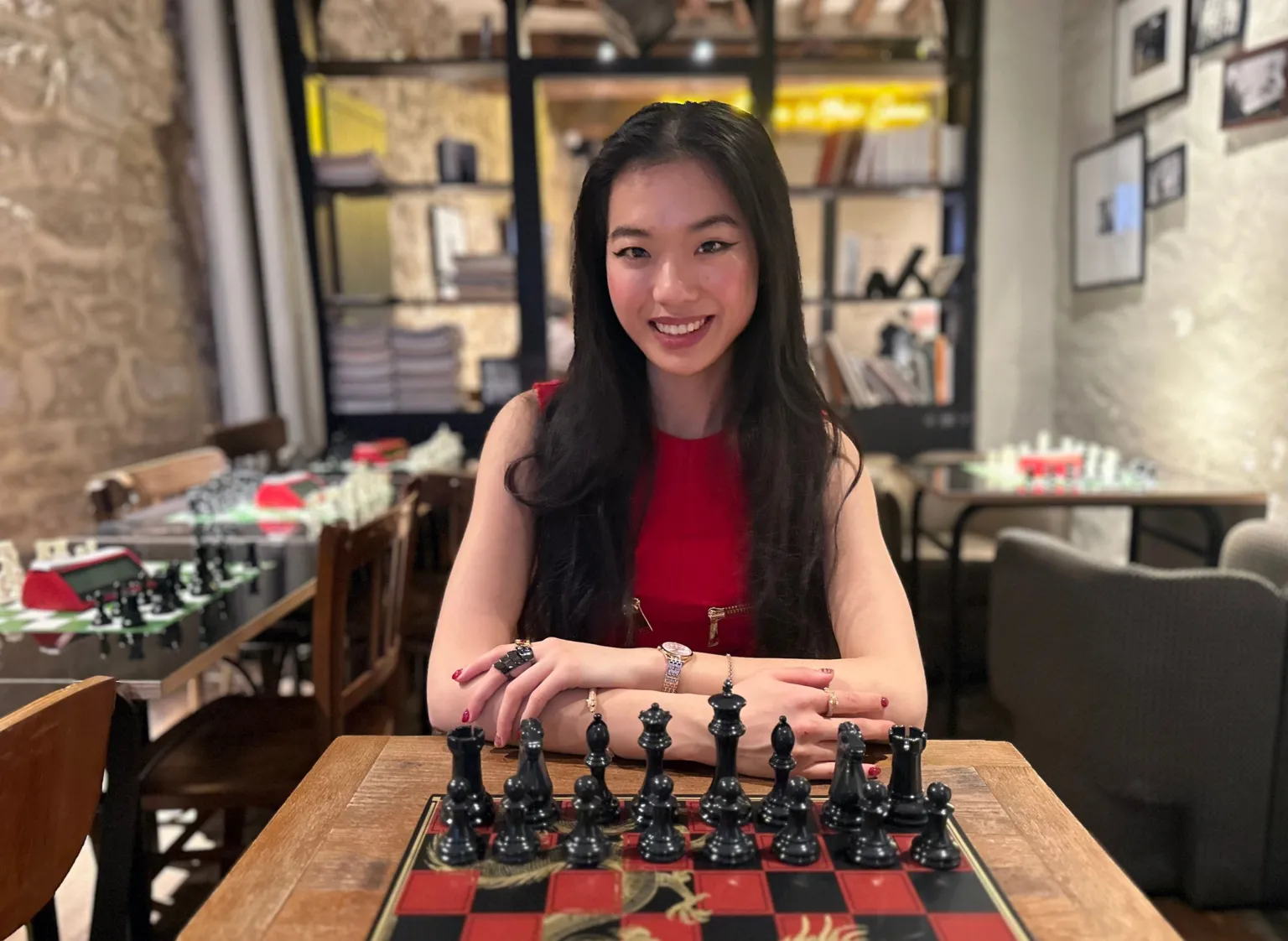 A young woman sitting in front of a chess board in an empty bar. She is wearing a red dress, her hair is dark long and straight. She is smiling at the camera and her hands are on the table before the board 