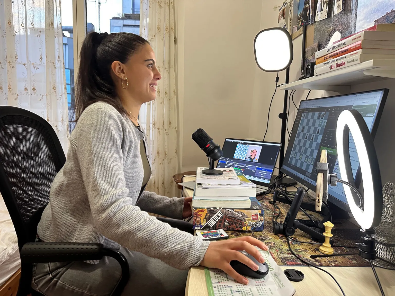 Sarah El Barbry is sat at her desk, smiling to the camera which is in front of her. On her desk there is a stack of books propping a microphone, a small ring light is on her right and there are two screens in front of her. She is in a small bedroom. 