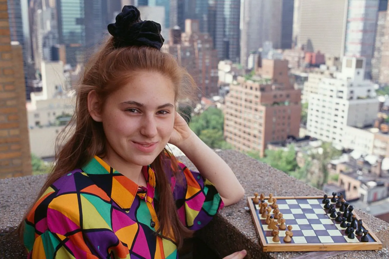 Sygma via Judit Polgar as a teenager pictured on a balcony of a high rise building in a big city. She is standing next to a small chess board, wearing a colourful shirt. Her hair is in a high ponytail with a large scrunchie. She is smiling at the camera.