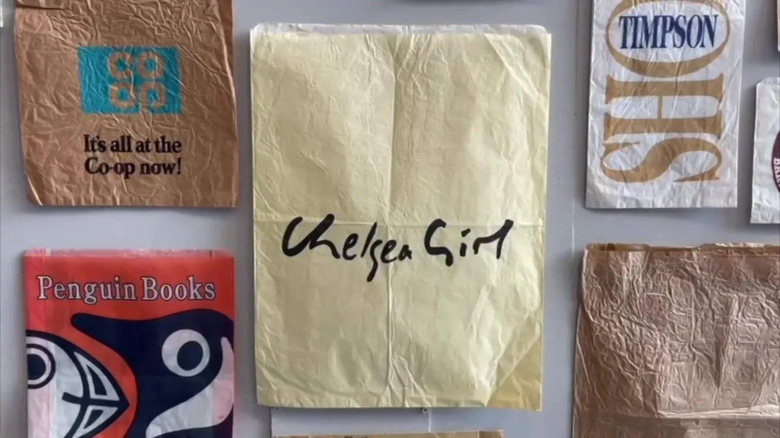 A collection of different paper bags pinned up on a wall as part of an exhibition. They include designs for retailers including Chelsea Girl, Penguin Books, and Co-op.