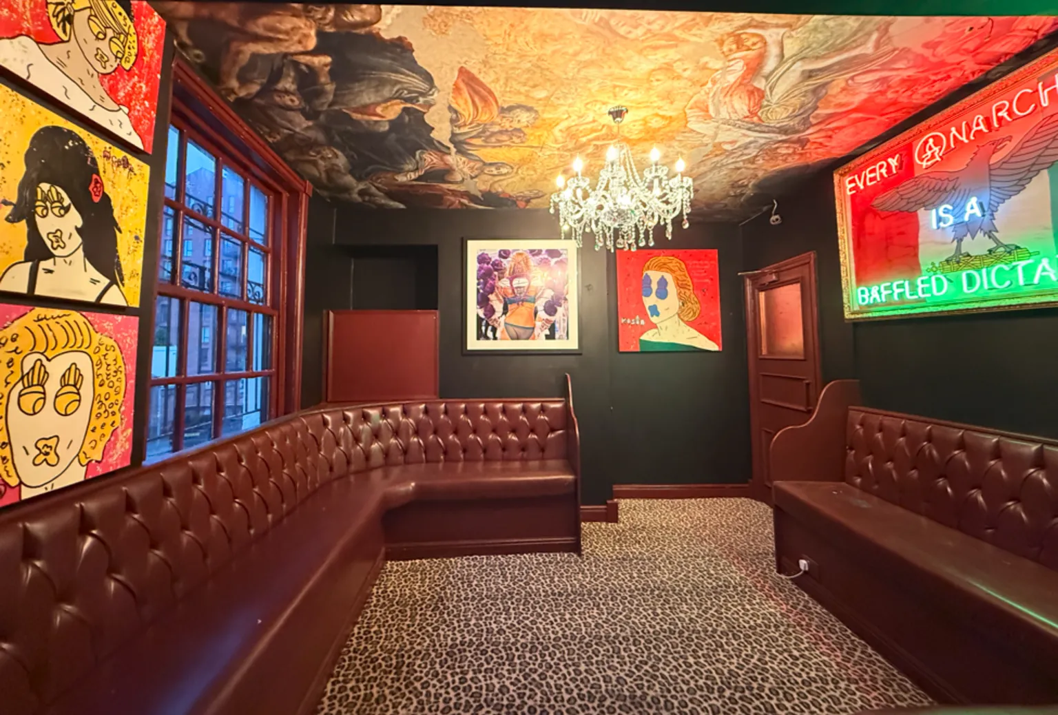 Mason Newman Leather settees surround two walls with vibrant painted pictured and light installations on the walls and a chandelier on the ceiling, which has a full artwork on it, with leopard print-style carpet.
