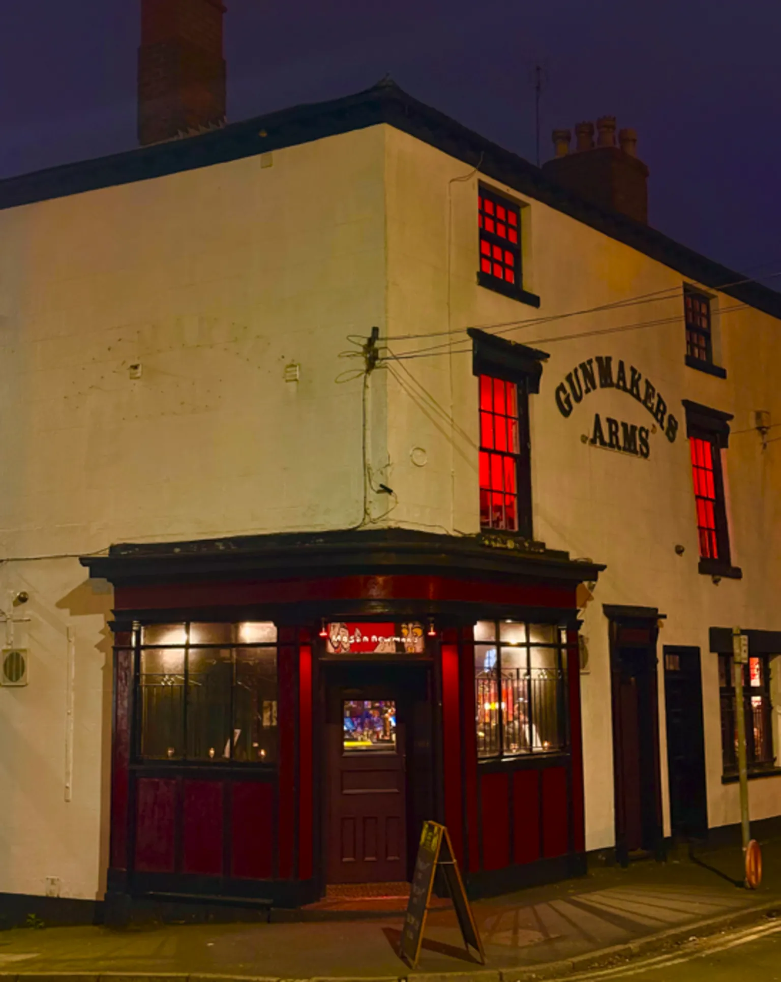 Mason Newman An external, angled view of the pub. It is white with red light glowing through the upstairs and downstairs windows. It has a wooden frontage around the door.