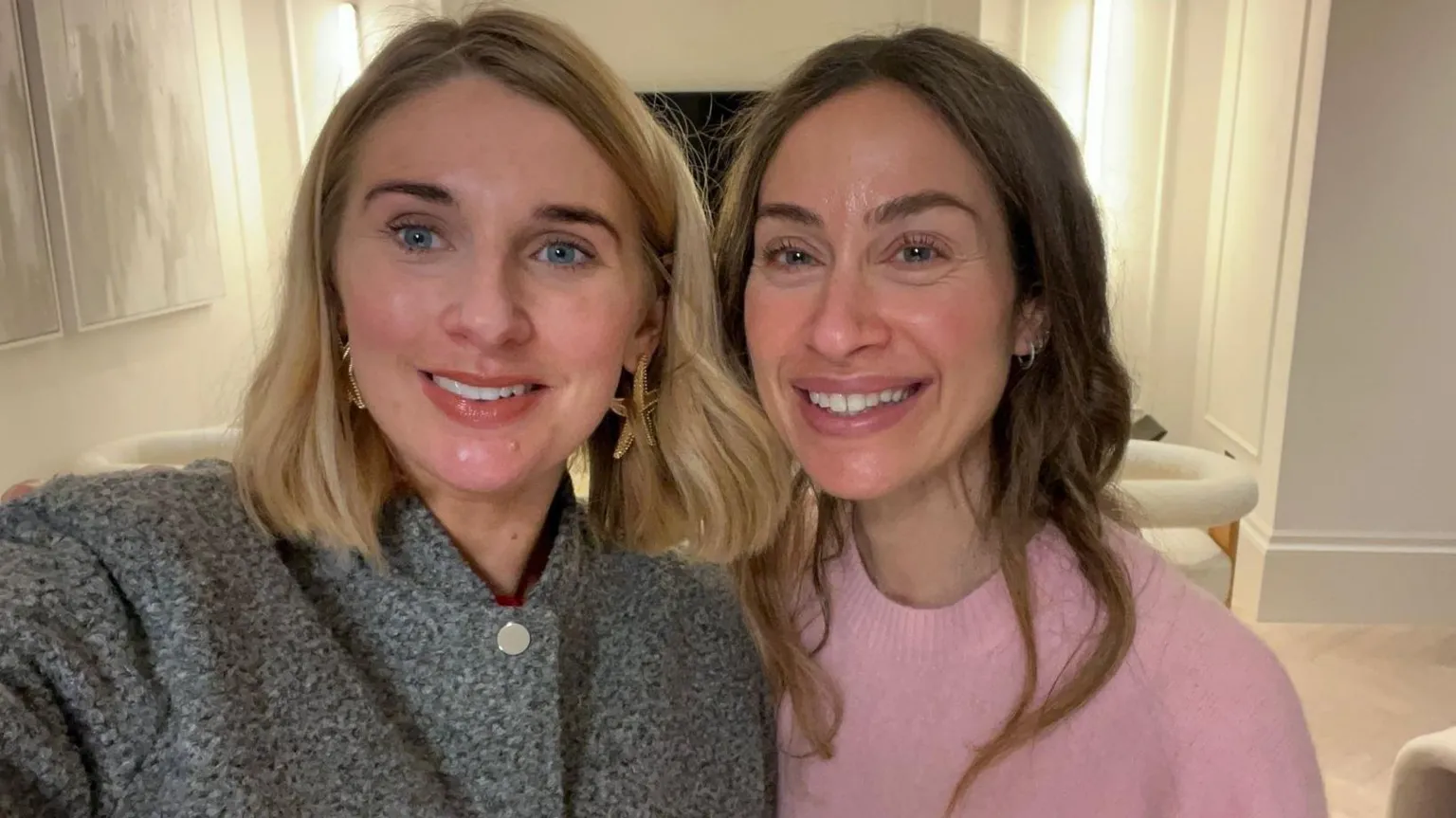 Two women smile into the camera, their heads together. the one on the left has shoulder length blonde hair and is wearing a dark grey top, the one on the right has long brown hair and is wearing a pale pink jumper.