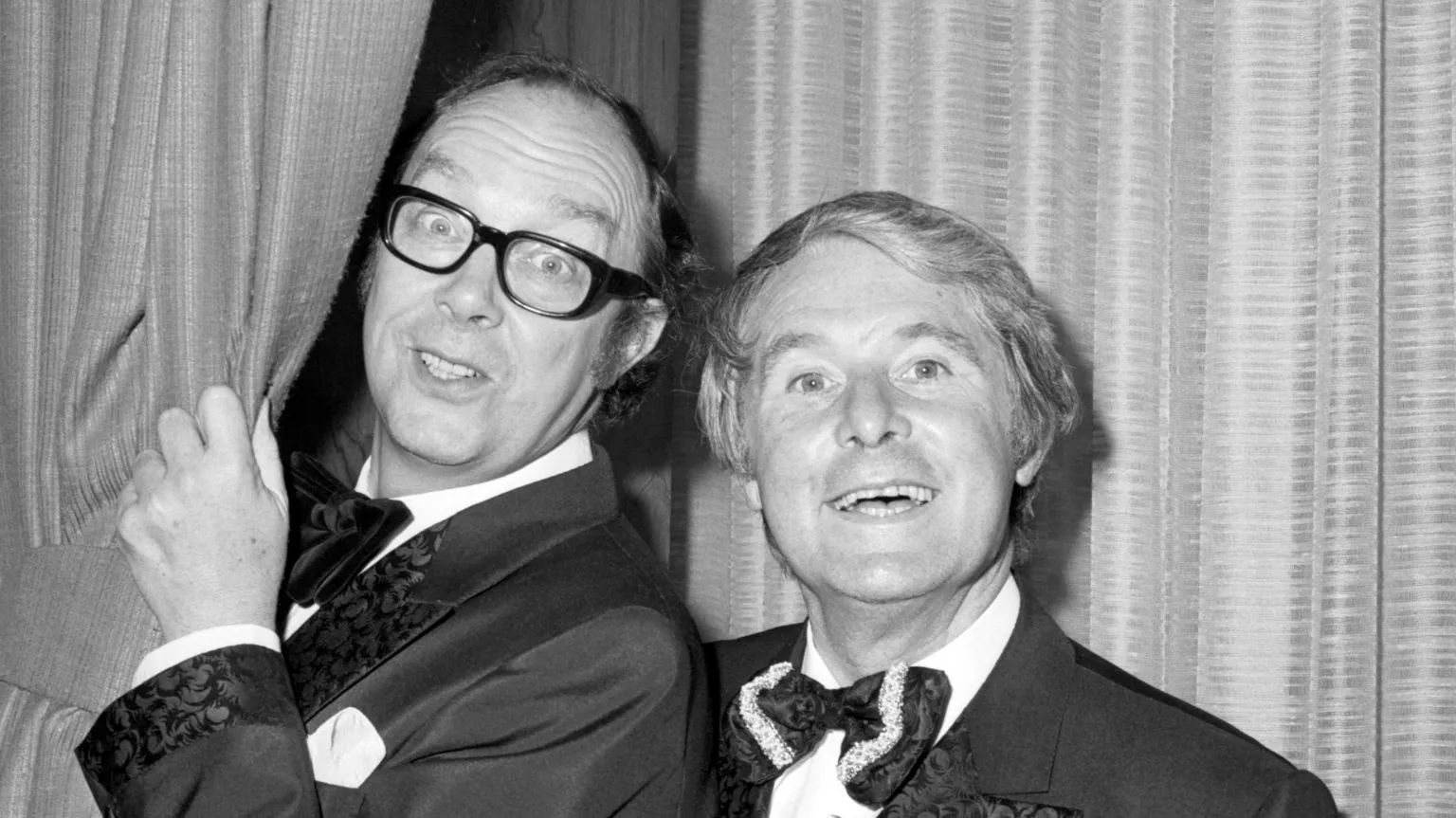  Black and white image of comedy duo Eric Morecambe (left) and Ernie Wise during one of their popular shows. Morecambe is holding a stage curtain. They are both smiling.