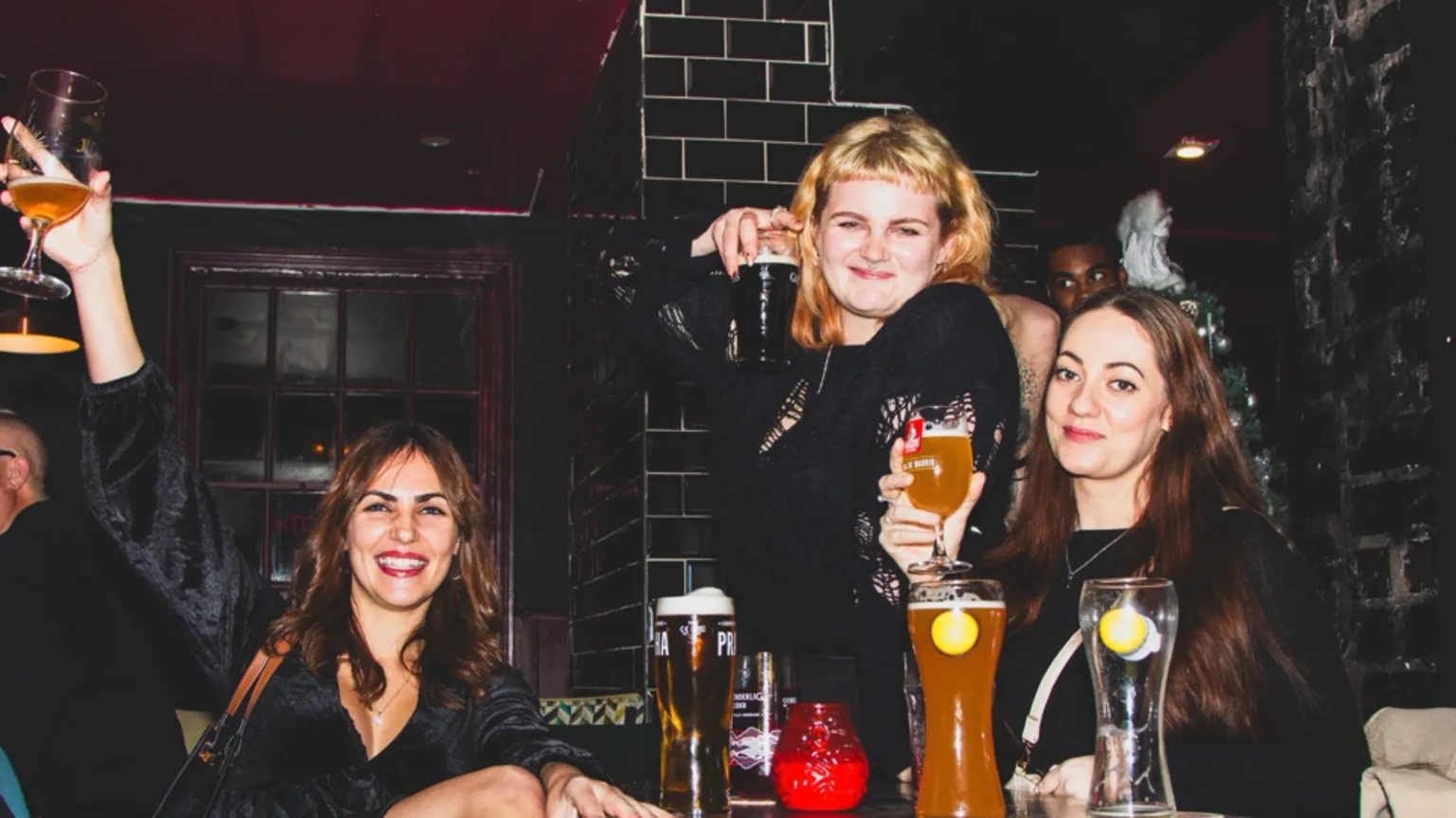 Mason Newman Three girls pose for a picture in the pub holding drinks while sat at a table smiling at the camera. One girl's arm is outstretched with a drink in her hand. The pub has a dark background with black tiles.
