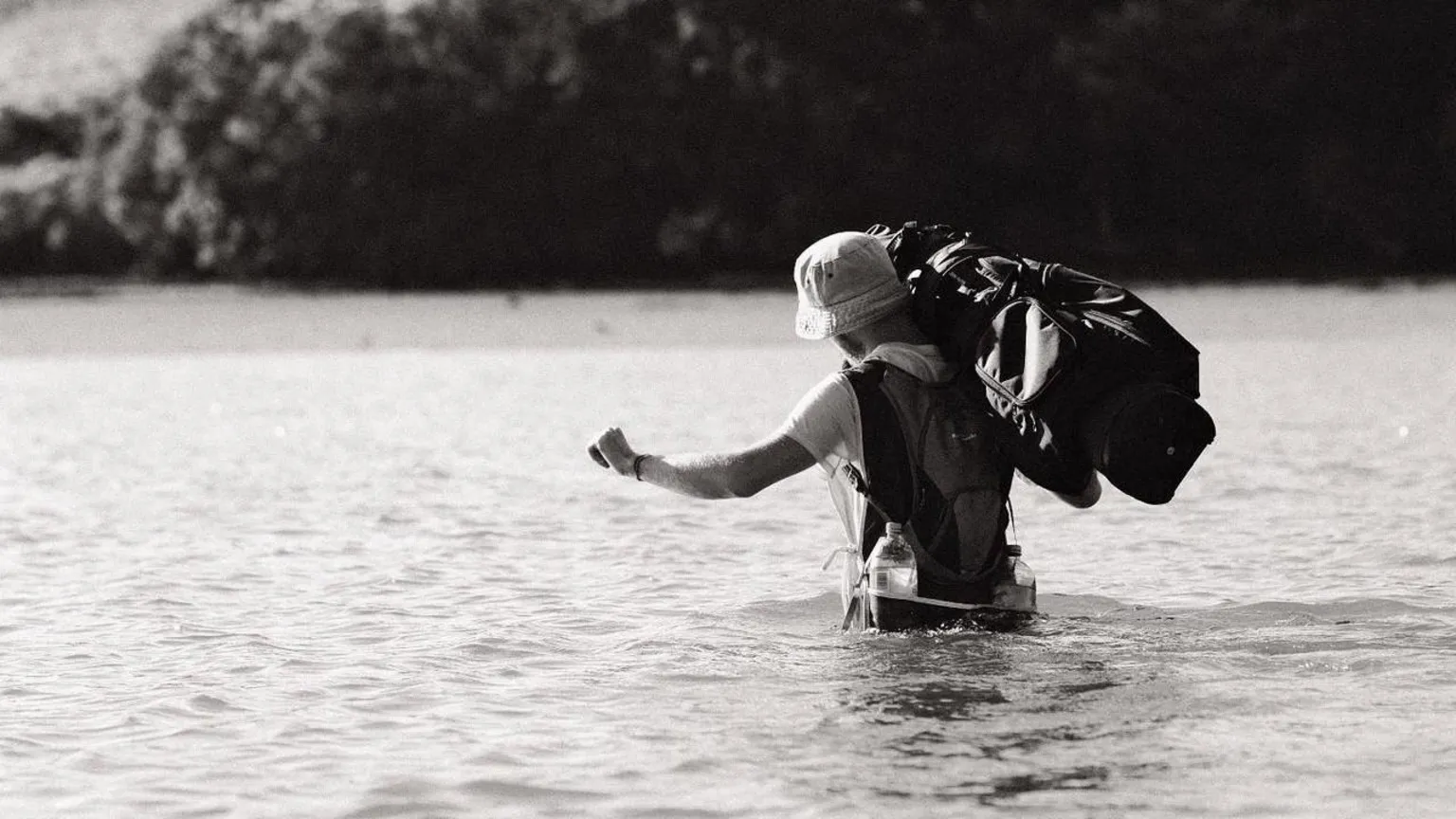 Dougie Haynes A black and white image of Dougie, who is wearing a bucket hat and t-shirt, wading through waist-deep water whilst carrying a golf bag on his shoulder.