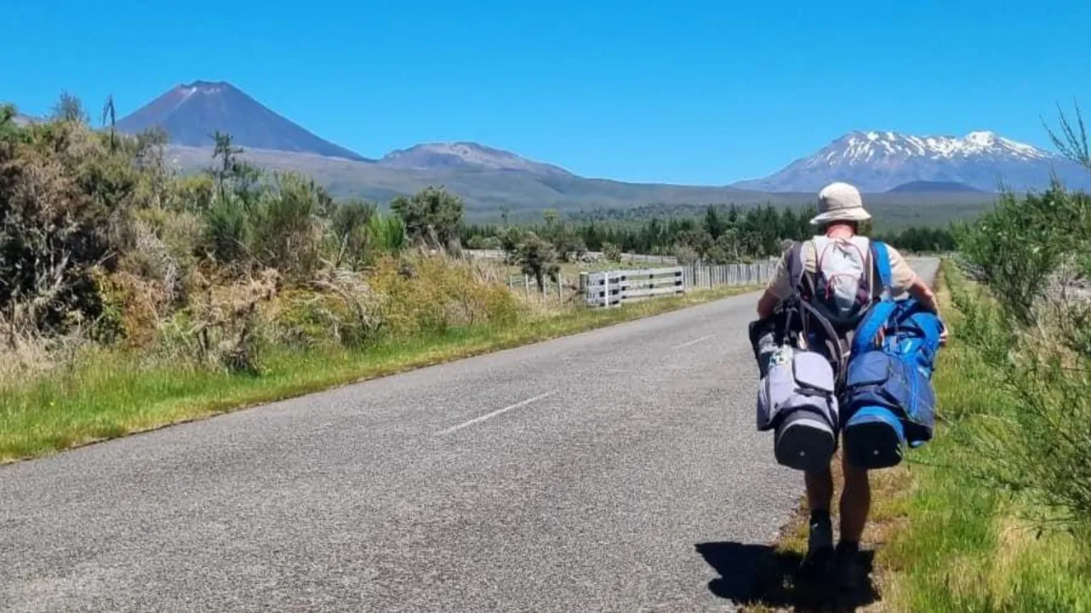 Dougie Haynes Dougie Haynes walks along a road carrying two golf bags. Two large mountains fill the horizon in front of him.