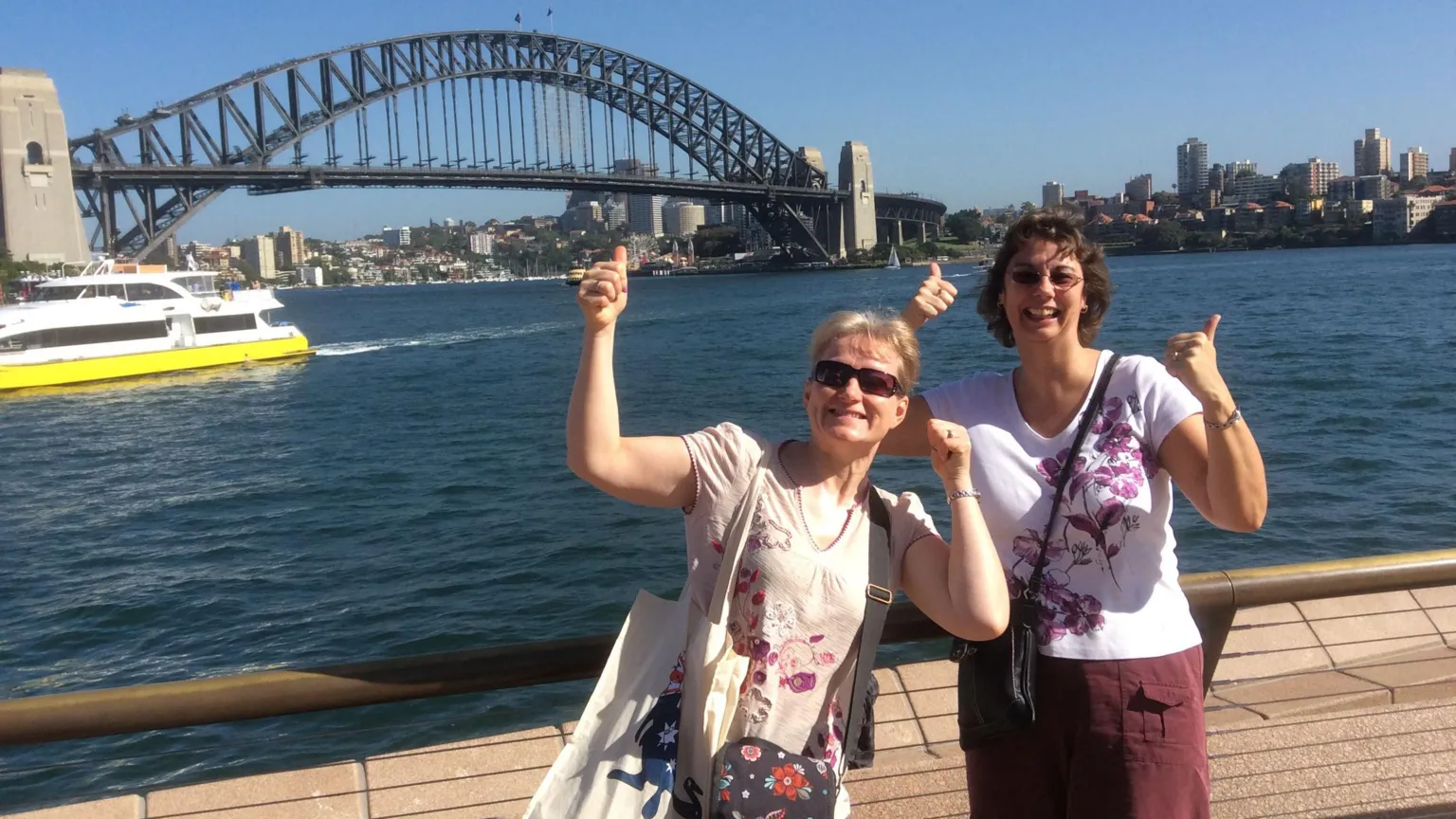 Ann Johnson Two women standing by the waterfront in Sydney, pointing upwards with the Sydney Harbour Bridge in the background.