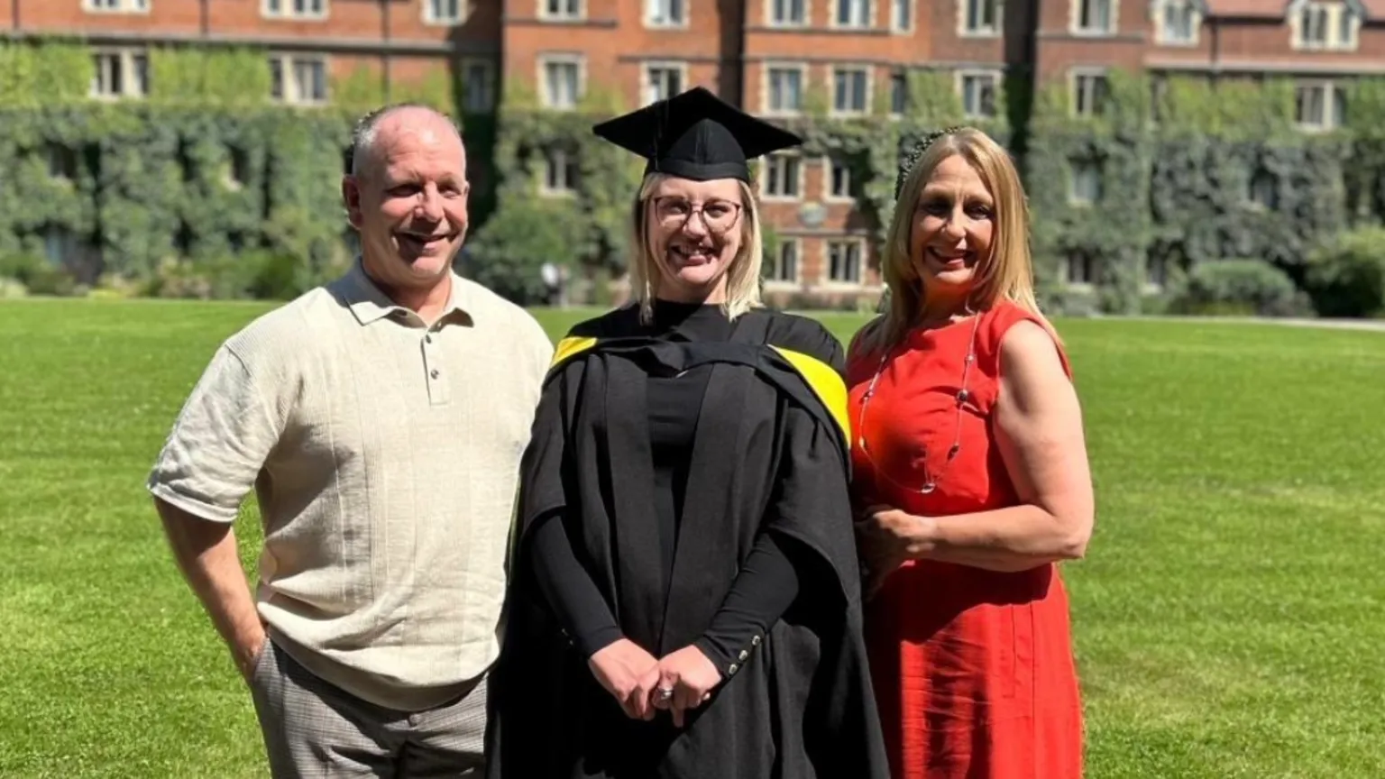 Handout Three people standing in front of a lawn of an old building. Charlotte's father on the left with little hair on his head and wearing a polo shirt. Charlotte in the middle wearing all black graduation robes including a mortarboard on her head. Joanna Donohoe on the right wearing a red dress. All three are looking at the camera on a sunny day, smiling.