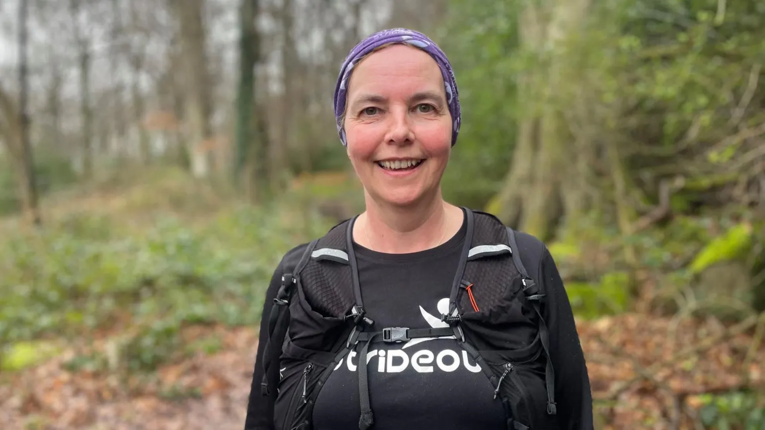 Simon Thake/BBC A woman in a black running top and purple head scarf smiles as she stands in woodland.