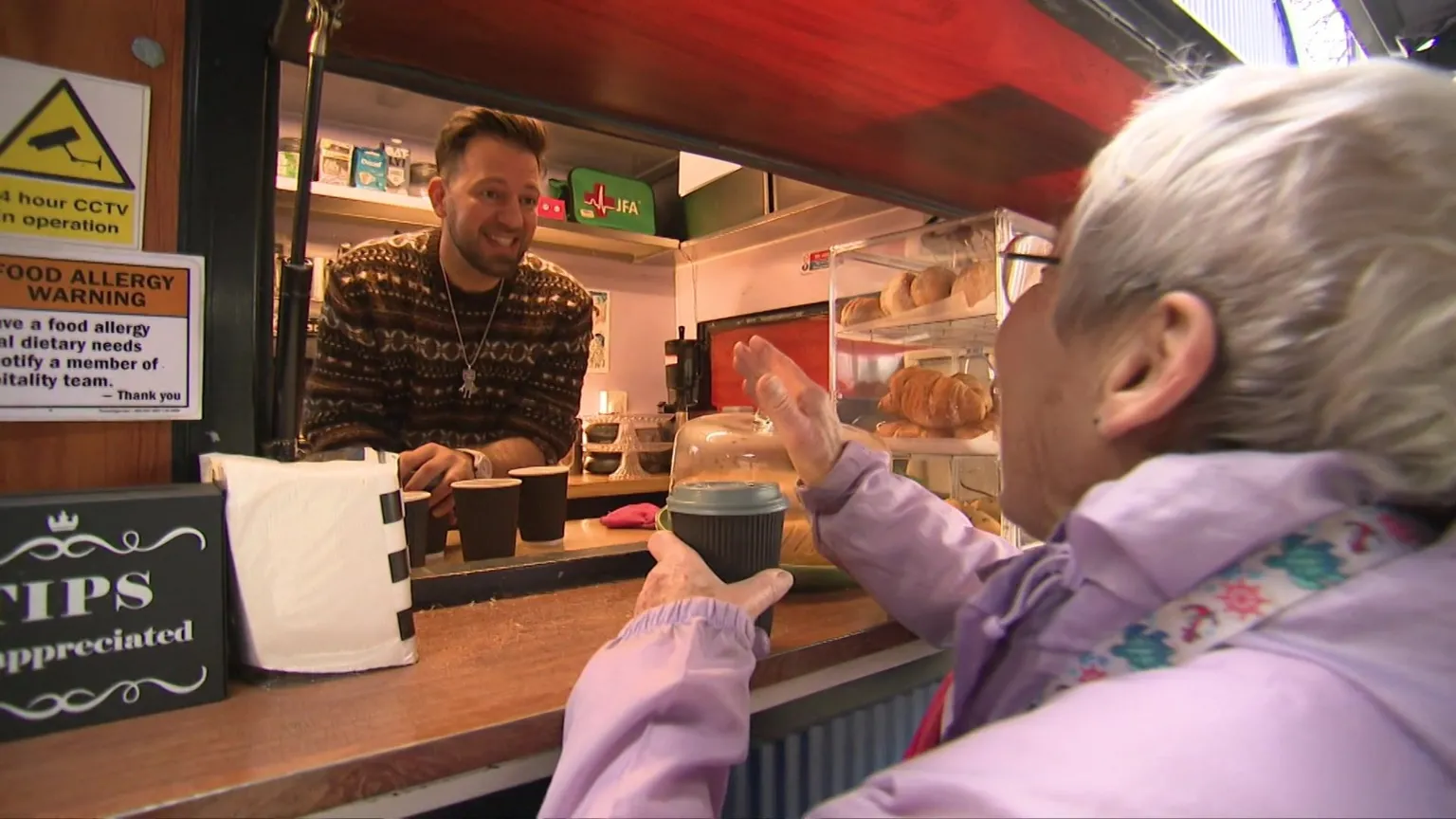 A woman in a pink jacket buys a coffee through the hatch in the coffee stand. She is smiling and waving, and Diaco is smiling back