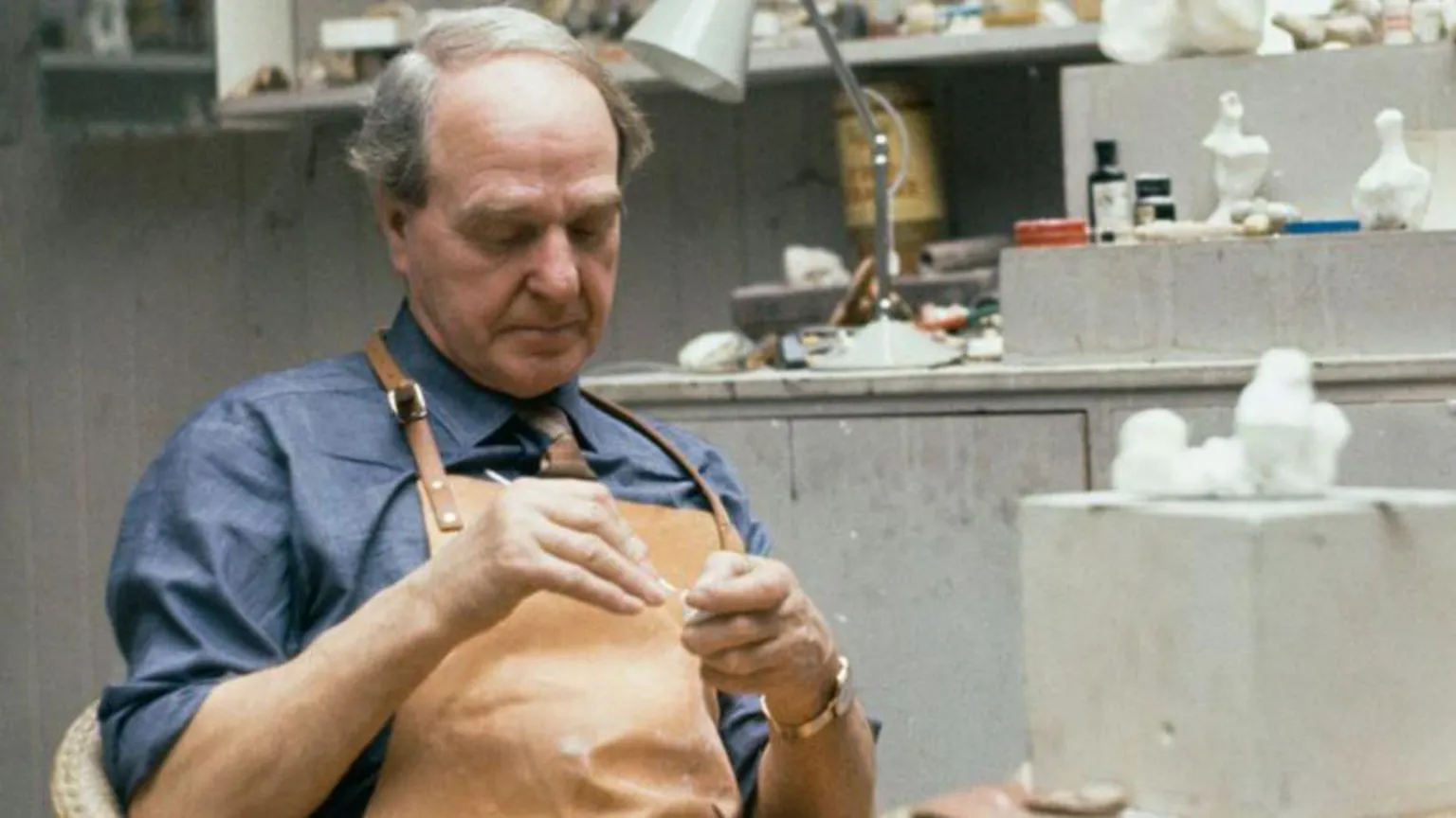  The artist Henry Moore in his studio, he is wearing a leather apron with a blue shirt underneath and has grey hair swept over his head. There are various artists' materials in the background.