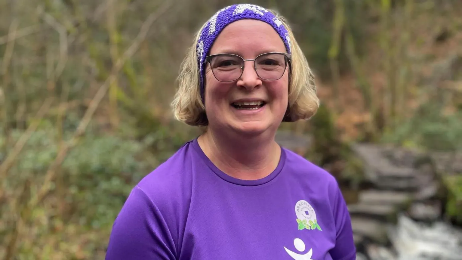 Simon Thake A woman in a purple running top and matching head scarf stands smiling in the woodland