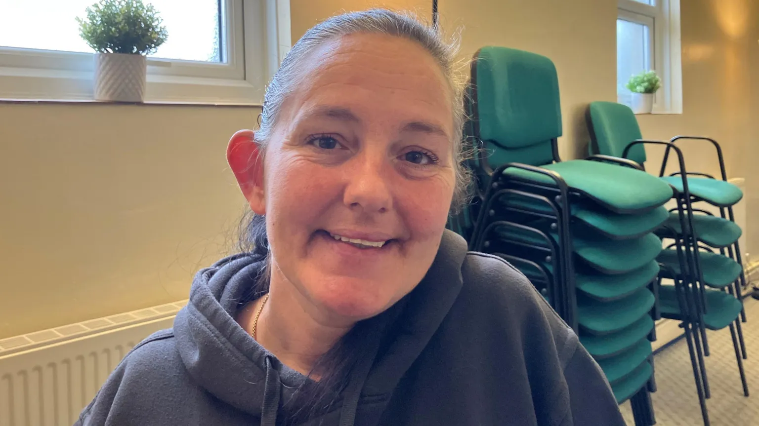A woman in her forties with greying hair in a ponytail, wearing a grey hoodie and sitting on a chair