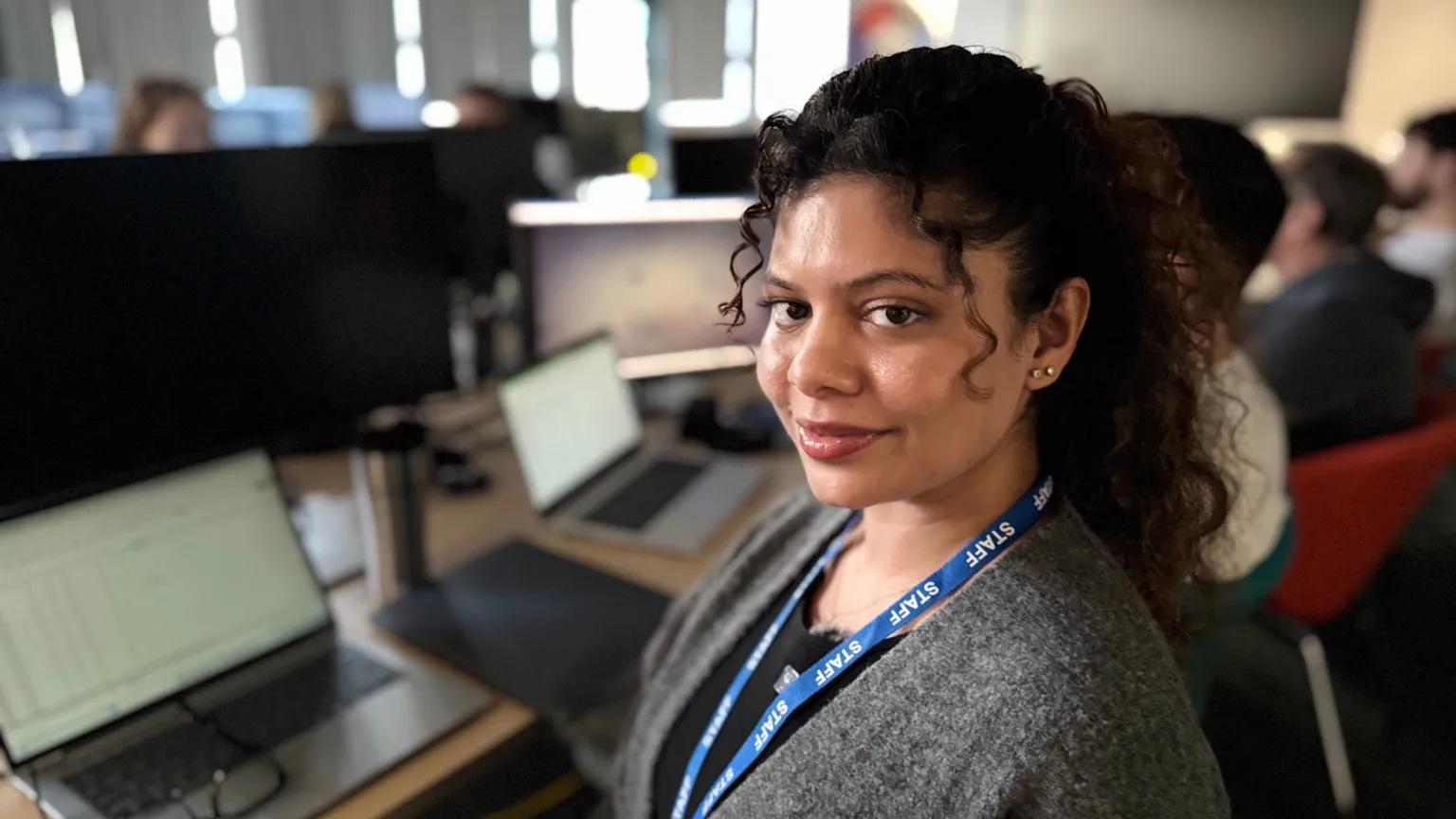 A woman with frizzy black hair looks to her left, into the camera. She sits at a desk where a grey laptop sits open in front of her. She wears a black t-shirt, with a grey cardigan and blue lanyard around her neck. Behind her are multiple desks with people sitting at them.