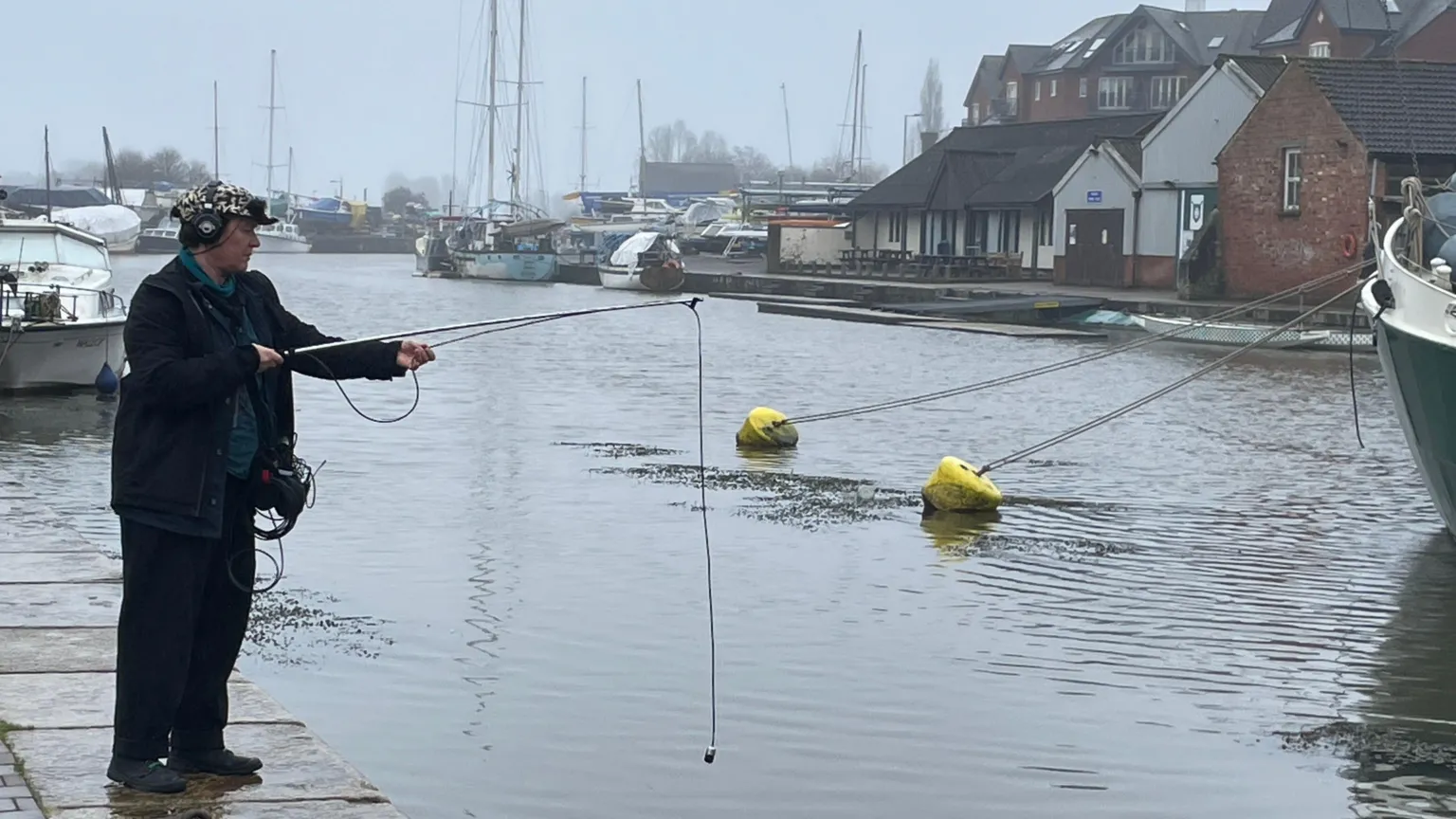 A woman dressed in dark clothing holding a microphone that is waterproof on a large stick, hovers it above the water. She is also wearing headphones and a cap covered in microphones.