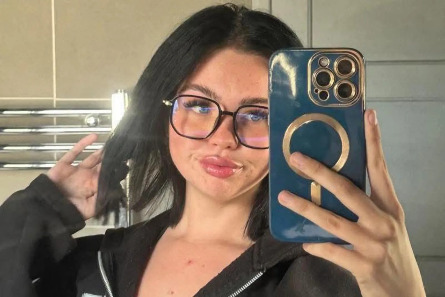 Supplied A woman with dark hair taking a selfie on an iPhone with a blue and gold phone case. She is wearing dark-framed glasses.