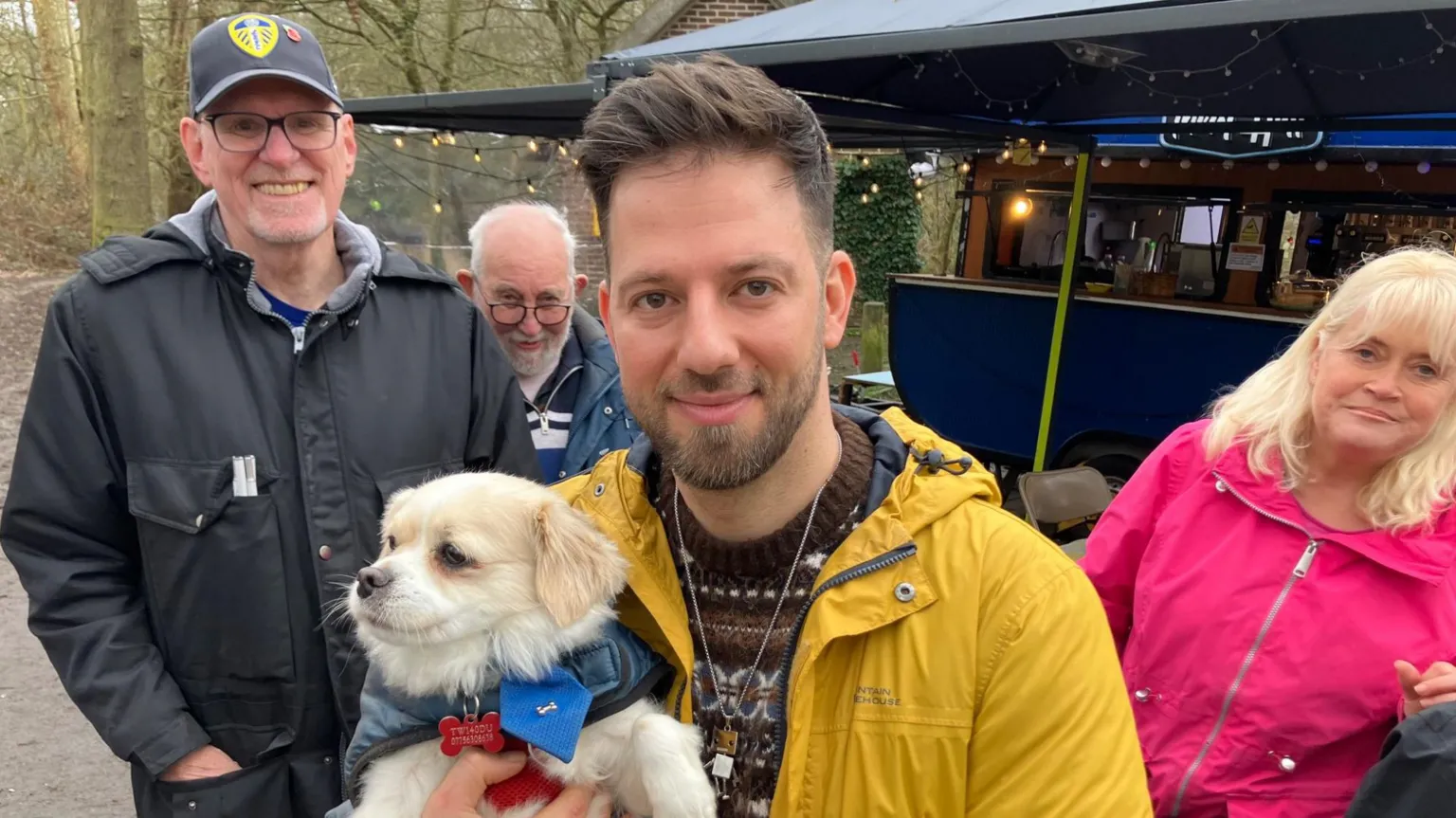Diaco is holding a small white dog and has a few of his customers nearby as they all smile at the camera 