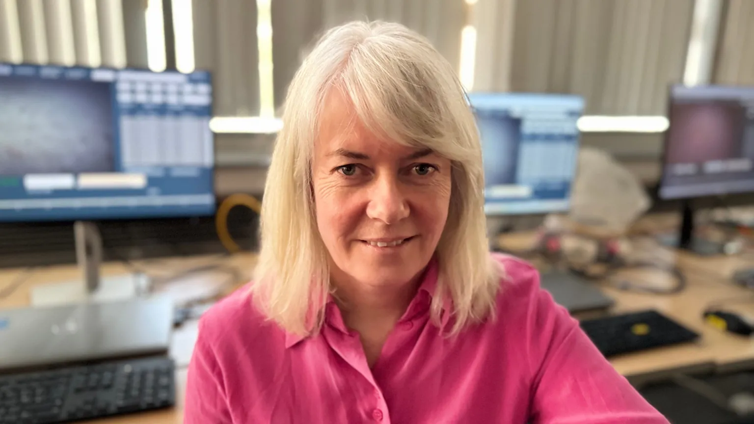 A long-blonde haired woman wears a pink shirt with an open collar. Behind her is a brown desk with three screens on it. She had brown eyes and smiles to the camera. The white blinds behind her are shut, they sit above the desks.