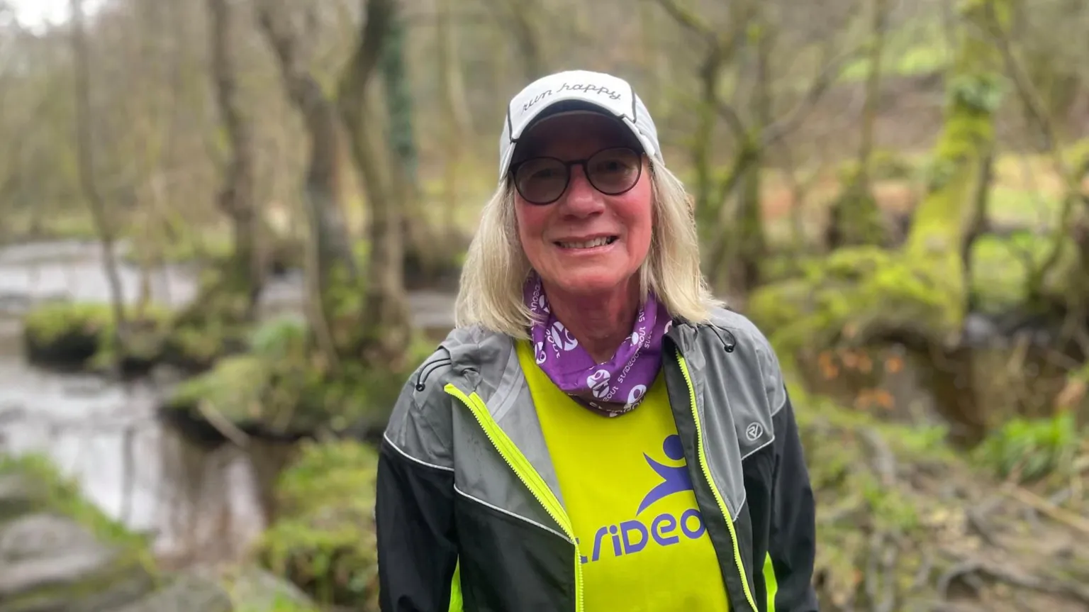 Simon Thake/BBC An elderly woman with long white hair in sports clothes and a silver cap stands surrounded by woodland.