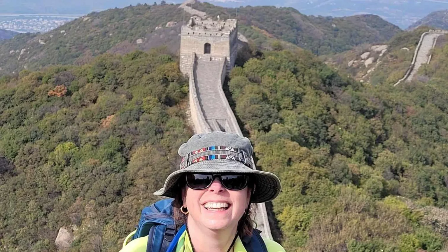 Cathie Rowe Cathie pictured with the Great Wall of China behind her. She is smiling at the camera and wearing sunglasses and a grey bucket hat.