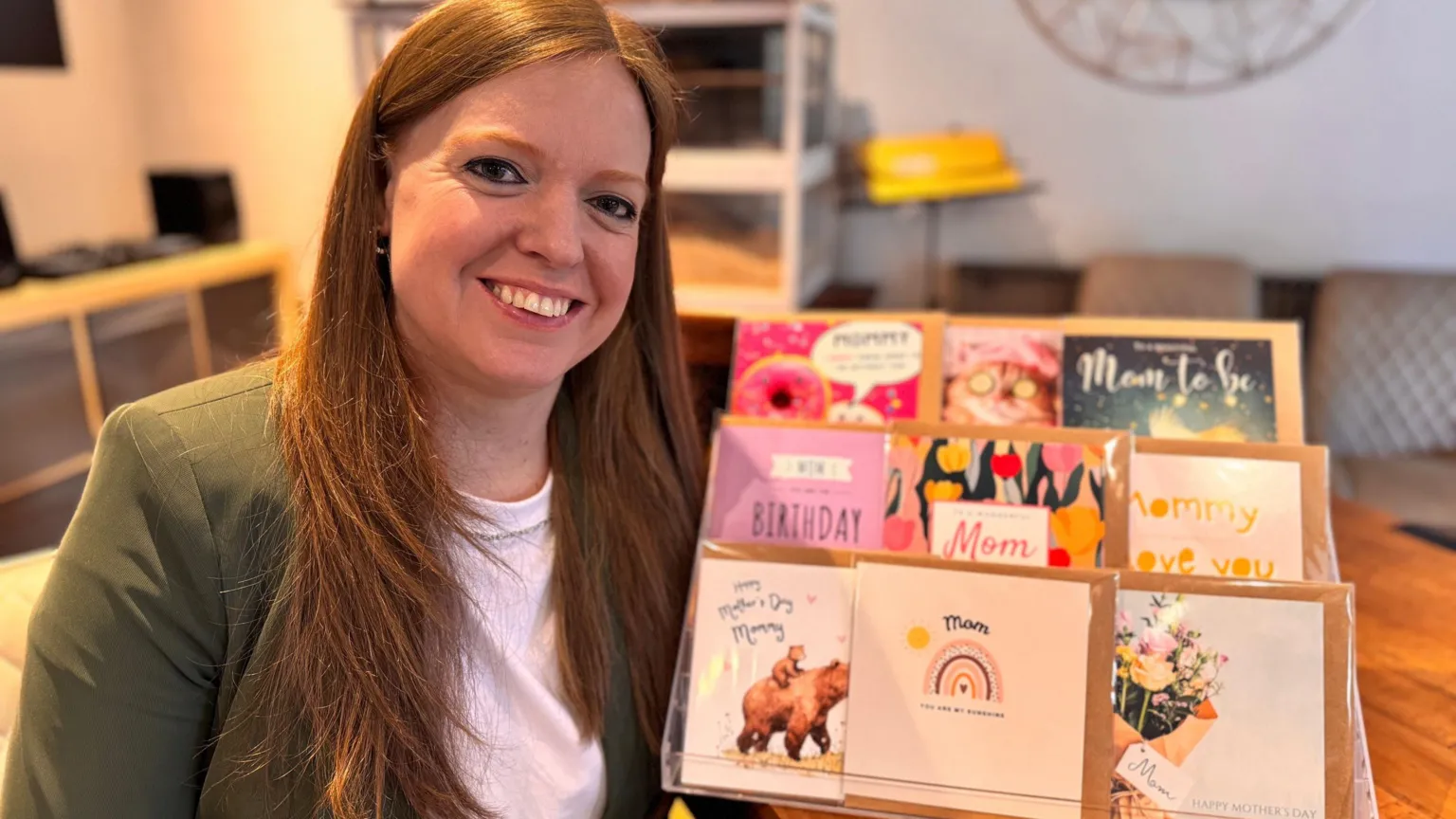 A smiling Andrea Pullen has dark red hair and is wearing a green jacket over a white T-shirt. Next to her on a desk is a stand displaying different birthday and Mother's Day cards.