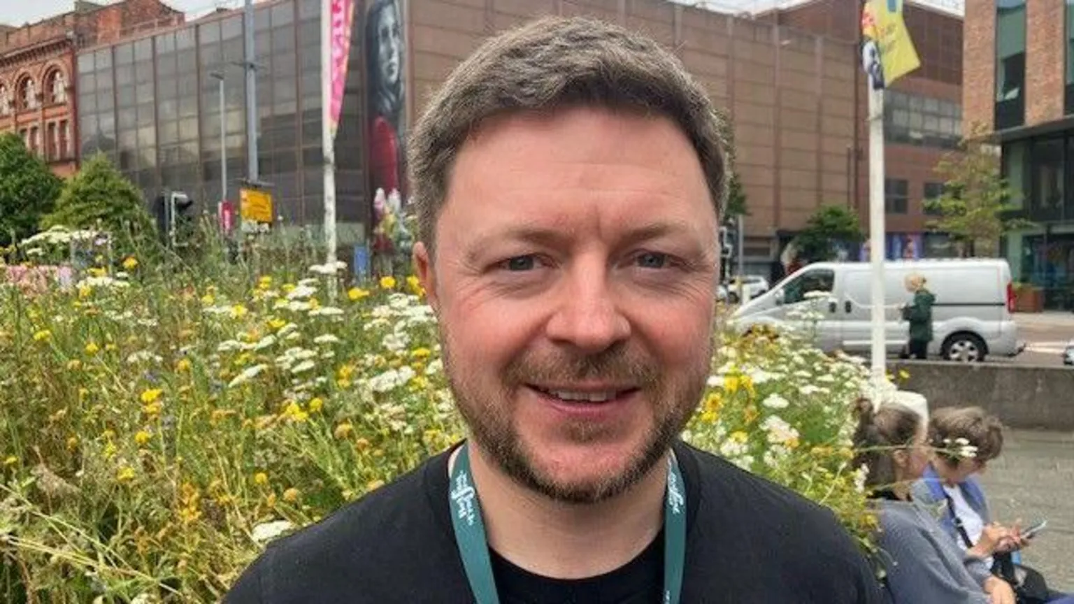 Dónal O'Connor. He has short brown hair and a beard is smiling, with wildflowers behind him as he stands in a city centre park.