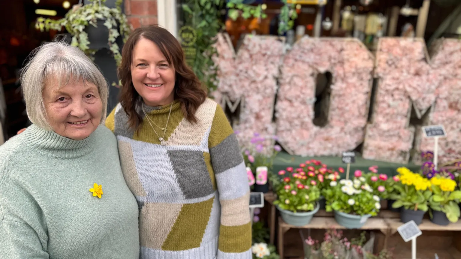 A smiling Penny Johnson stands in front of her flower shop with a floral display behind. She is wearing a grey, brown and white jumper and is standing next to her mom who is in a light green jumper.