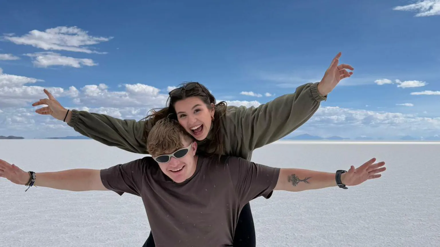 Sioned Cray Sioned is pictured on Fin's back. They are both waving their arms in the air. They are pictured at the Uyuni Salt Flats in Bolivia. Fin is wearing sunglasses and they are both smiling.
