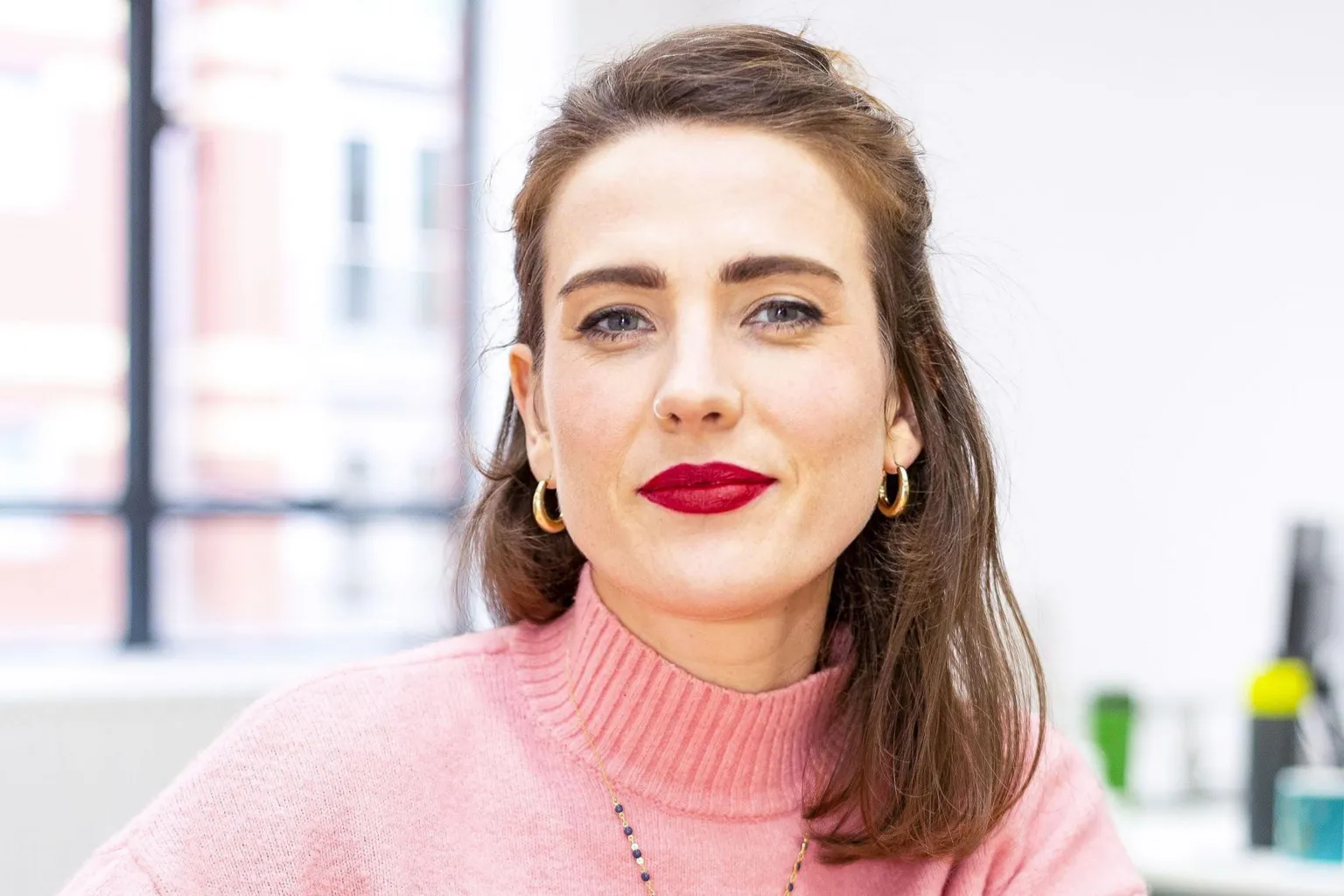 End Violence Against Women A woman in a pink jumper. She has brown hair and is wearing gold earrings.