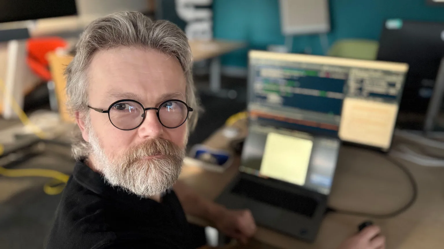 A man with black rimmed clear spectacles turns his head round to look at the camera. He sits at a desk with a grey laptop connected to a larger screen, via a black wire. The man has a grey beard and short grey hair.