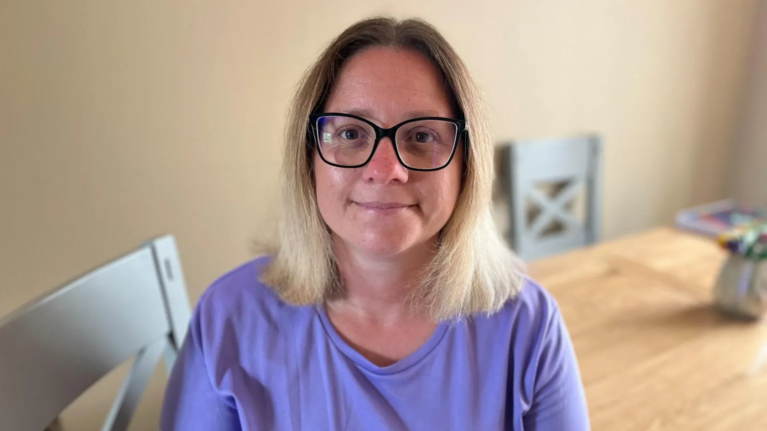 Qays Najm/BBC A woman with blonde hair wears black-framed glasses and looks at the camera. She is wearing a lilac top and sitting on a kitchen chair against a plain background.