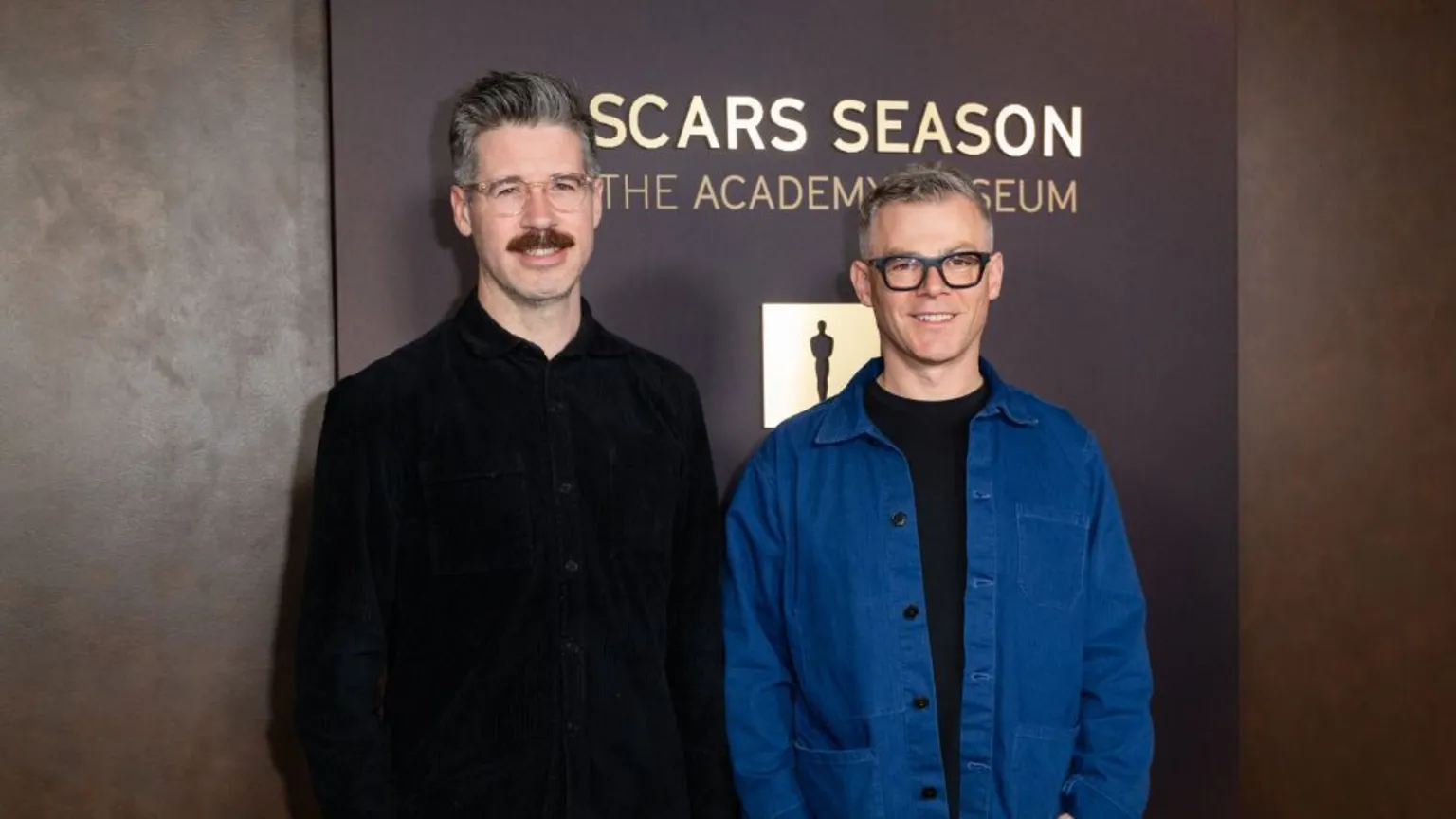 Two men stand side by side in front of a backdrop featuring the words “OSCARS SEASON” and “THE ACADEMY MUSEUM.” The man on the left is wearing a dark, button‑up jacket, and he has hair grey, glasses and a moustache. The man on the right also has short grey hair, wears glasses, and is wearing a blue jacket over a dark shirt. The backdrop is in warm brown tones with an Oscars statuette icon displayed between the text elements.