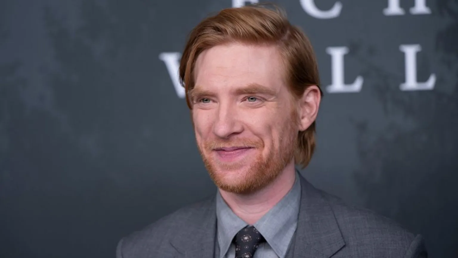 Actor Domhnall Gleeson wearing a grey suit jacket, light grey dress shirt, and patterned tie stands in front of a dark backdrop with large, partially visible white letters. He has ginger hair styled neatly, and the background features a textured, mottled design.