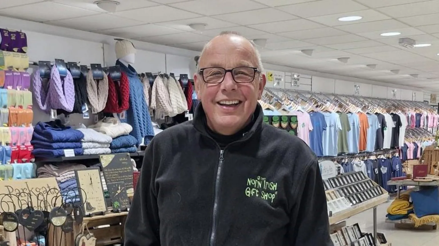 Keith Lane Keith Lane. He is bald, wearing glasses and a black zip-up fleece with green writing saying 'The Norn Irish Gift Shop'. Behind is wracks of t-shirts and scarfs and folded knits.