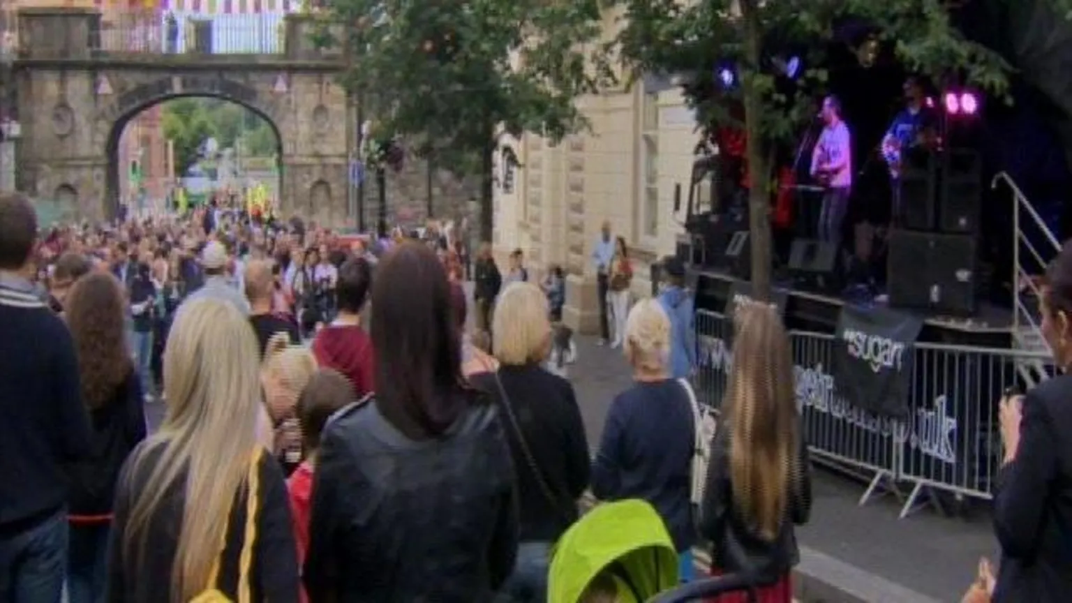 The back view of crowds gathered on a street. To the right there is a stage with a band playing. At the bottom of the street there is a stone archway.